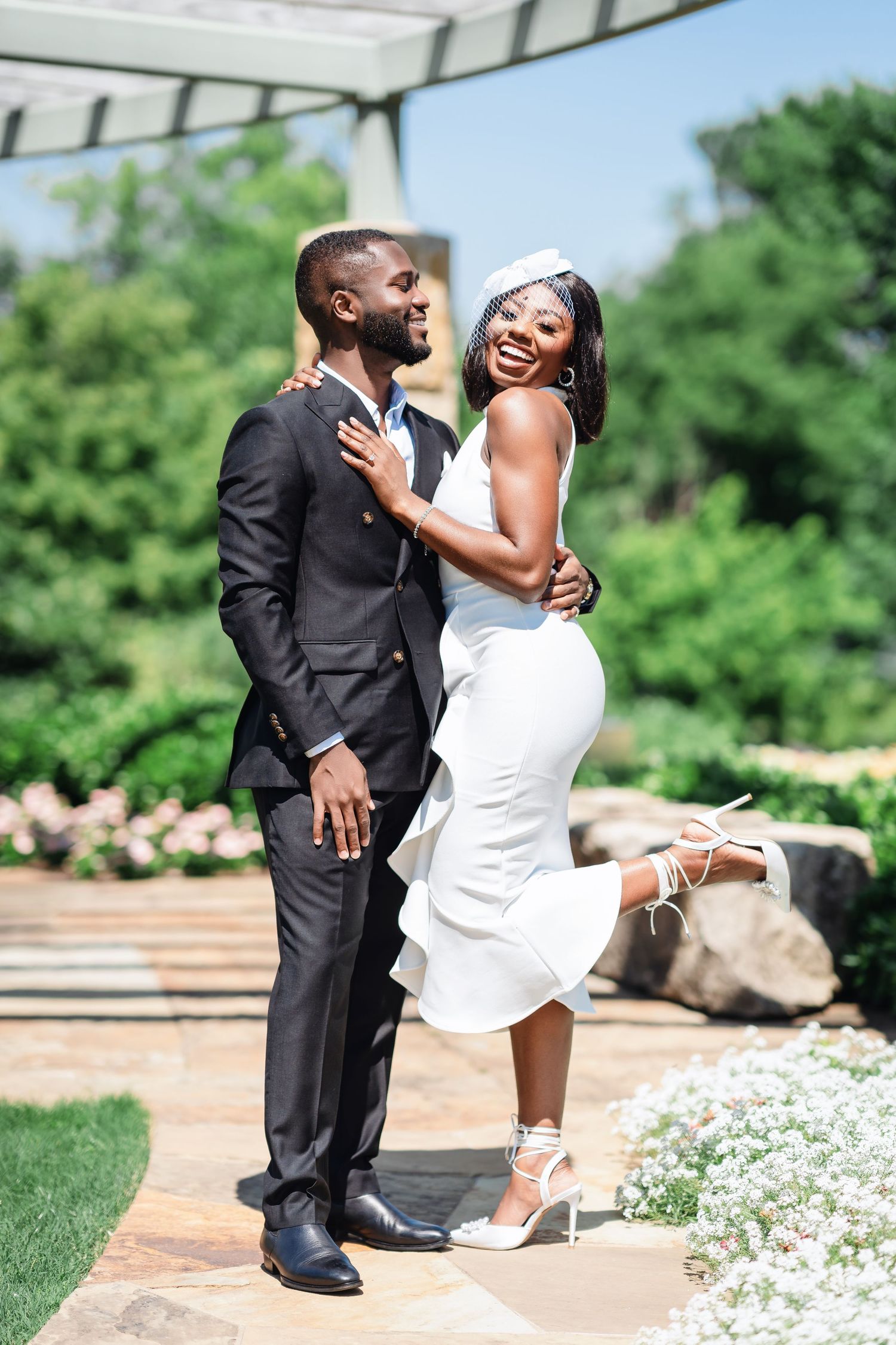 A couple shares a playful moment on a stone path surrounded by white flowers and greenery.