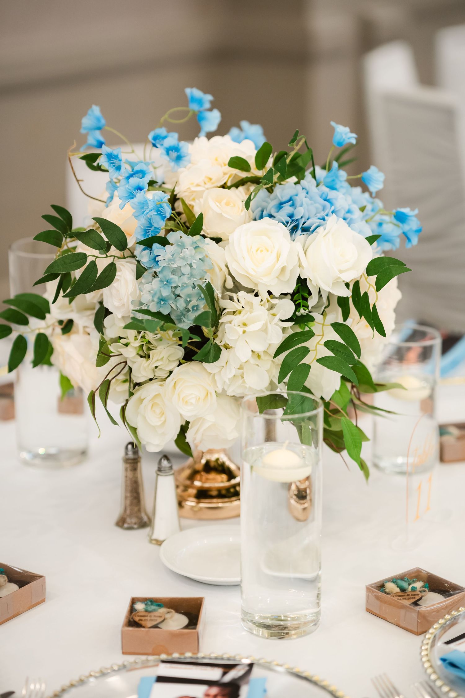 Elegant white and blue floral centerpiece with roses, hydrangeas and delicate greenery in a clear vase.