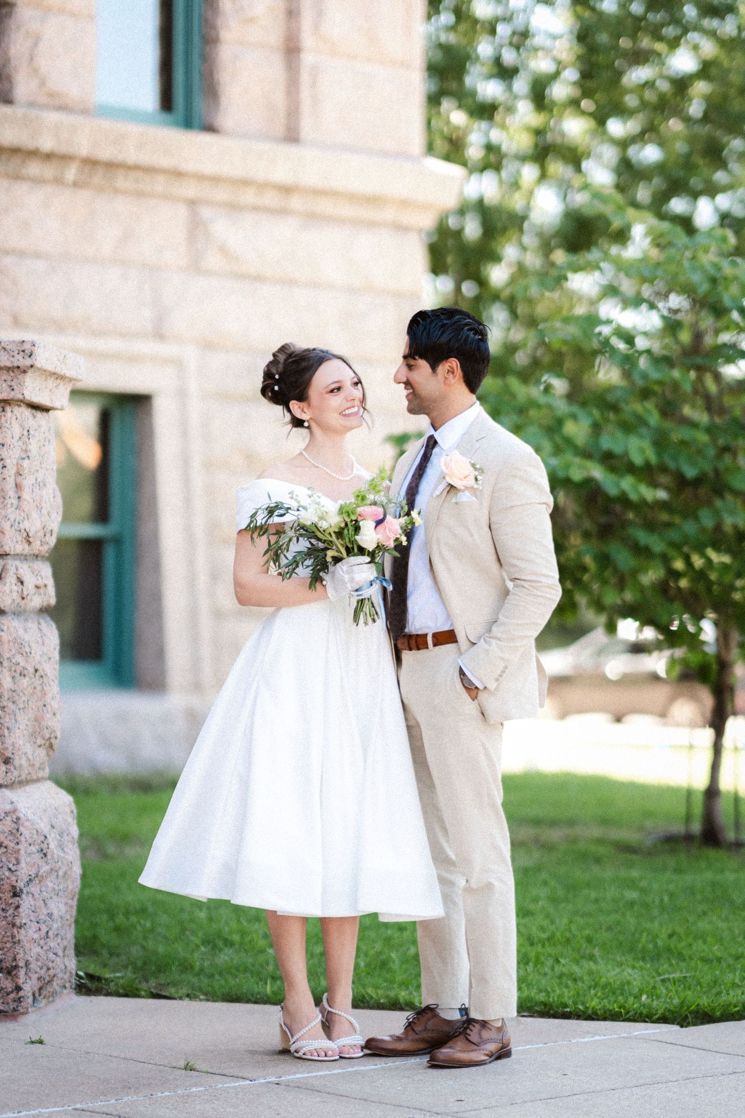 Wedding couple in white dress and beige suit pose playfully in front of a stone building with green landscaping.