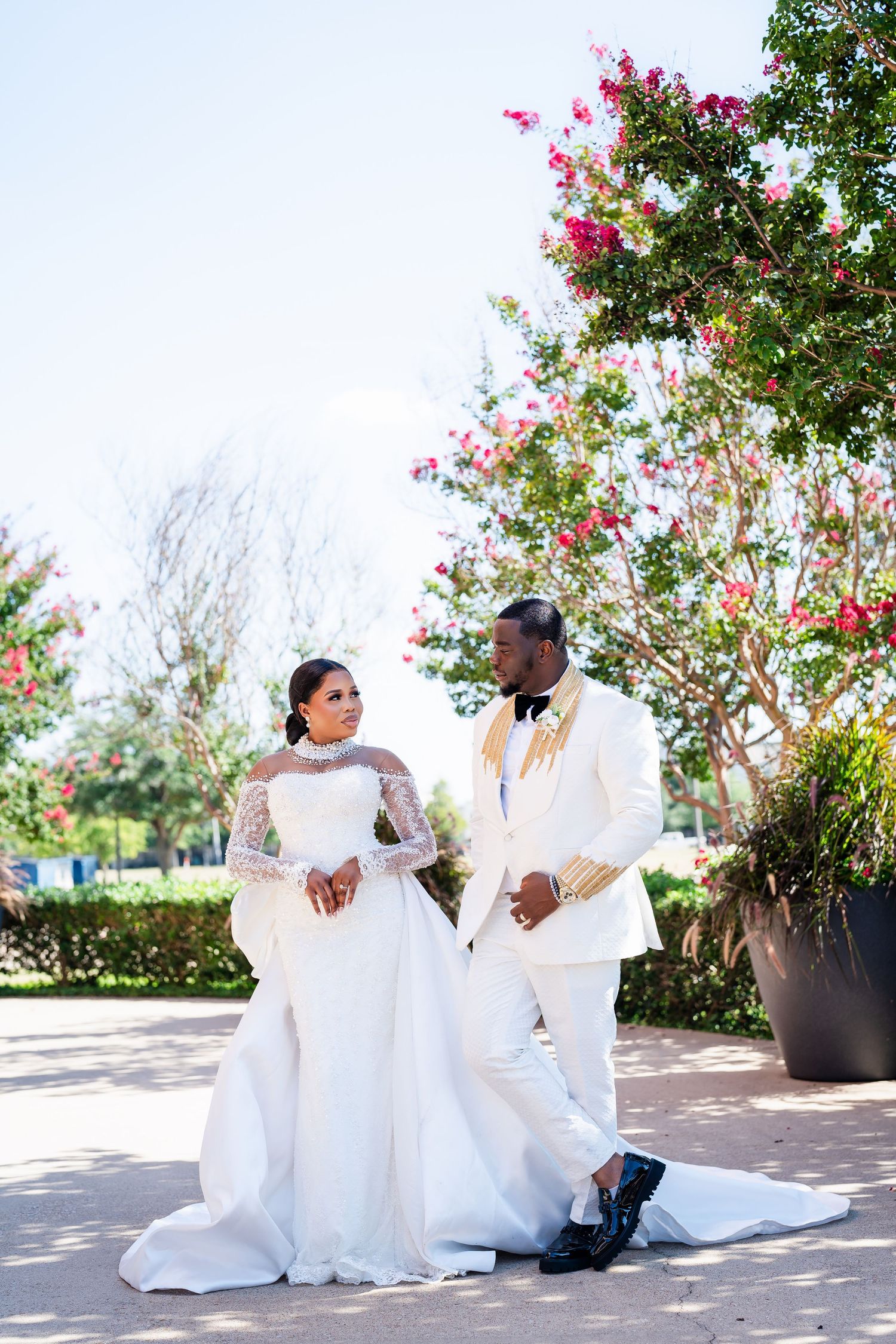 A couple in white wedding attire sitting together outdoors near blooming pink flowers.