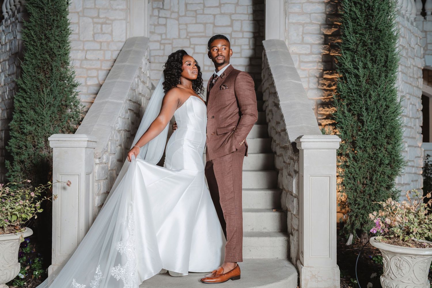 A couple in wedding attire pose on elegant stone steps between cypress trees at dusk.