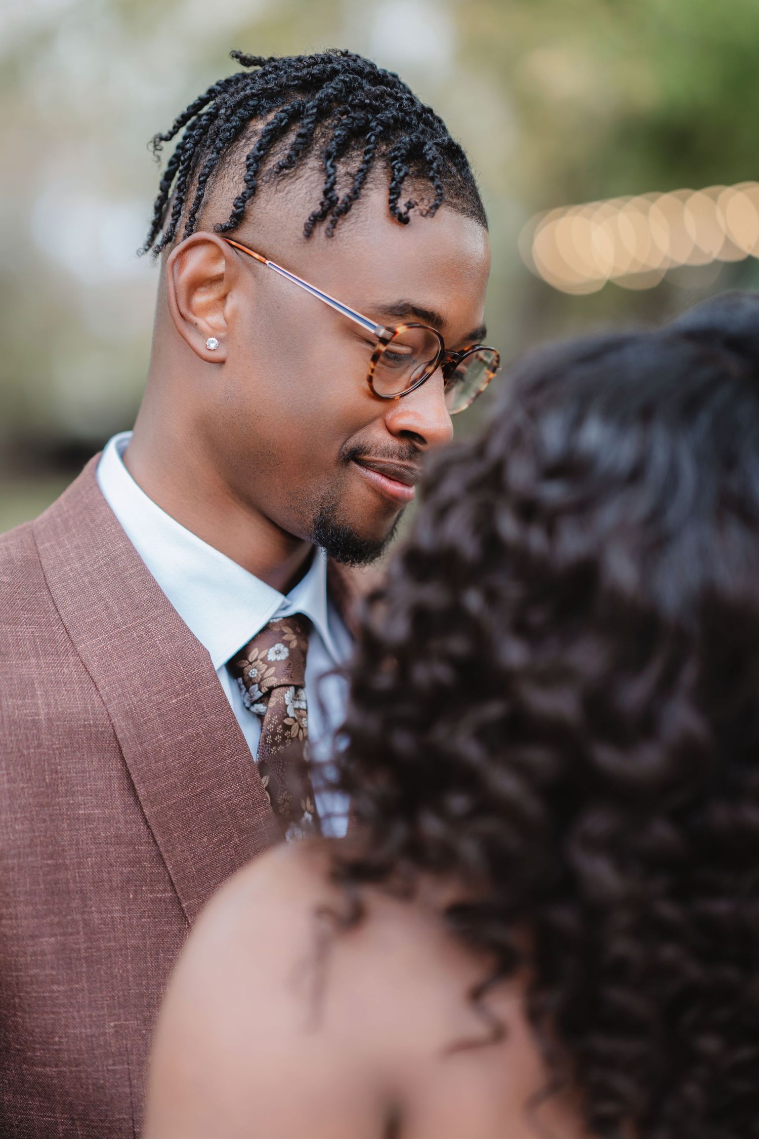 A groom wearing a brown suit and glasses smiles during an outdoor wedding ceremony.