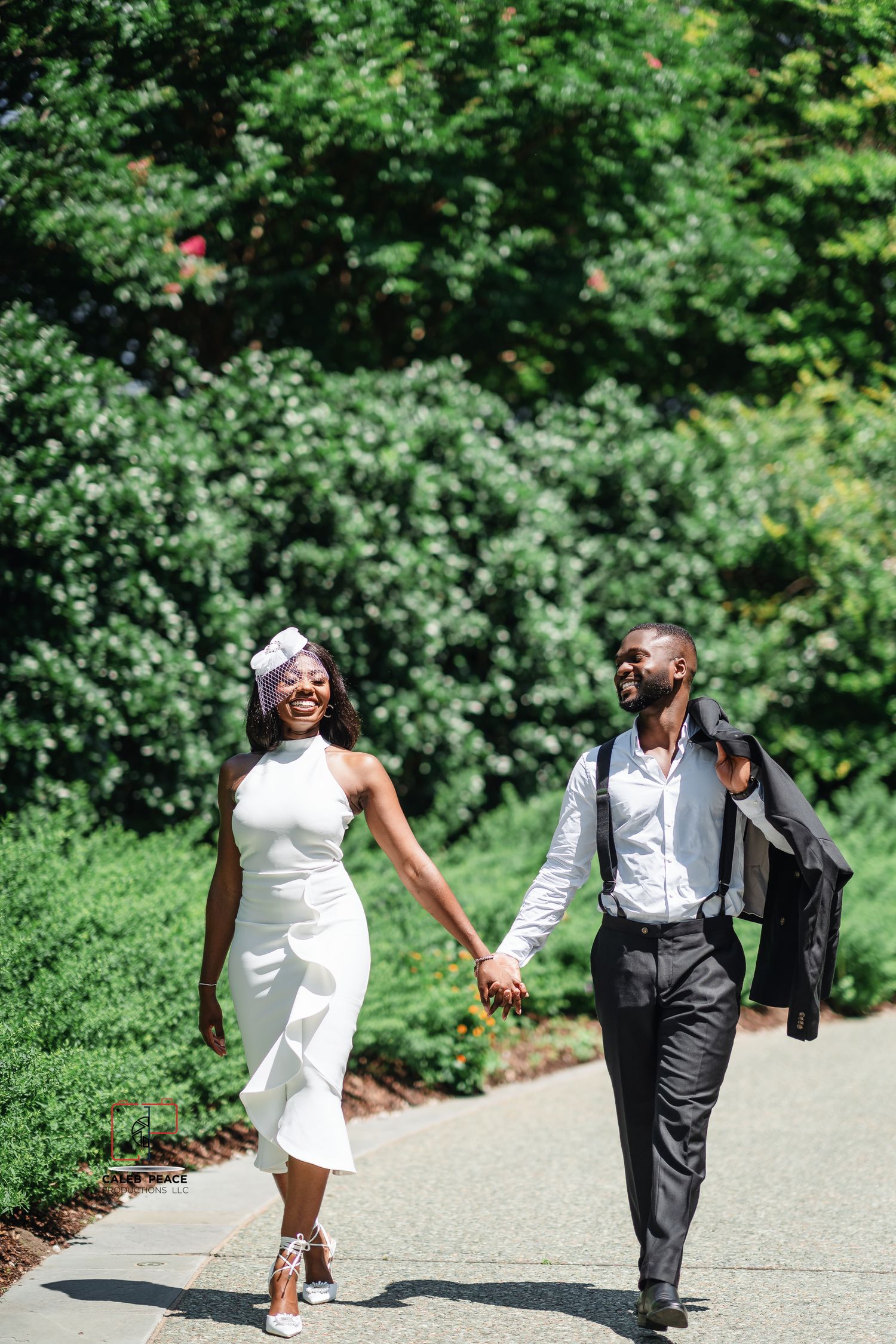 Couple walking hand in hand on garden path, wearing elegant white dress and dark suit on sunny summer day.