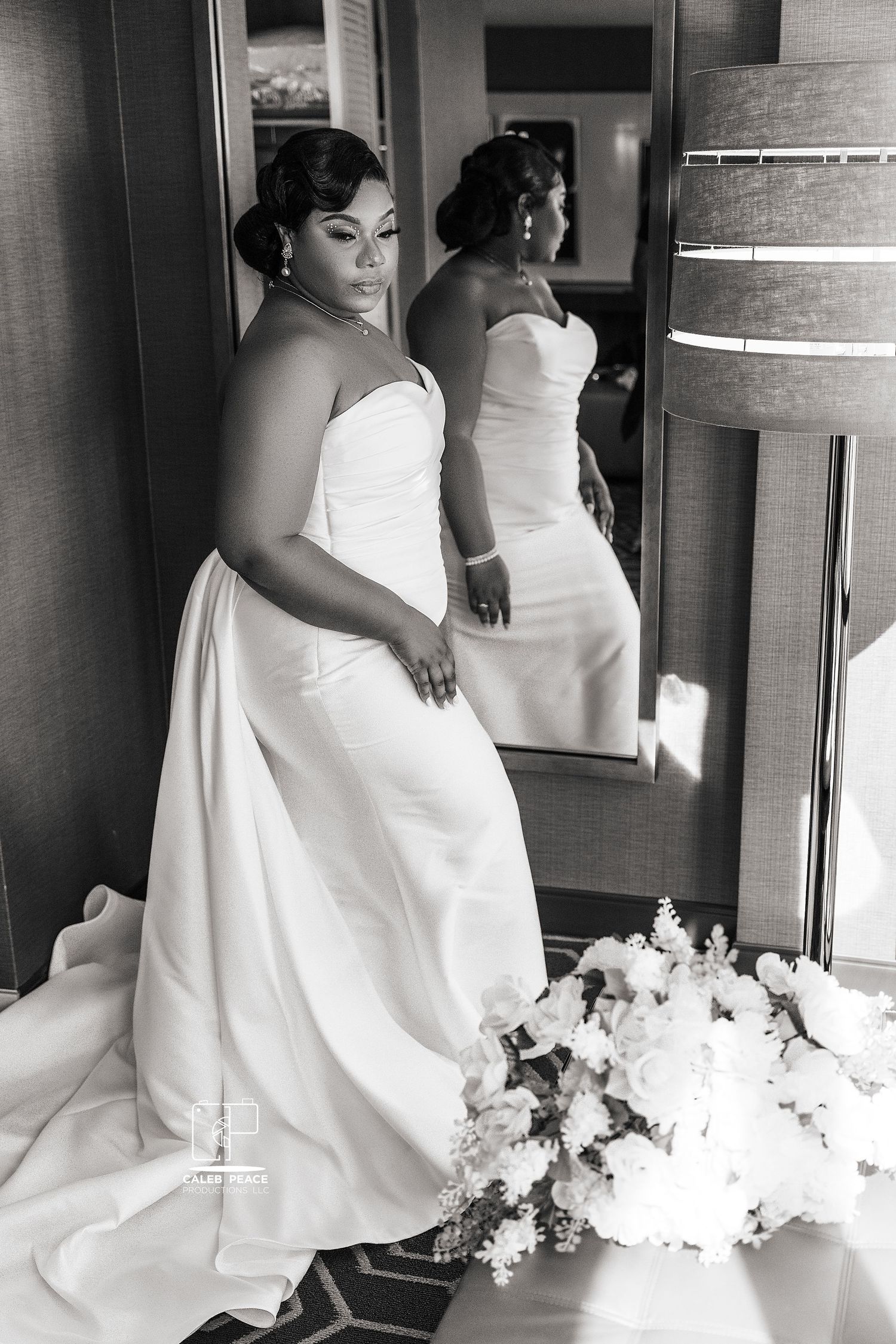 A bride in a white strapless wedding gown poses elegantly beside a mirror with a floral bouquet.