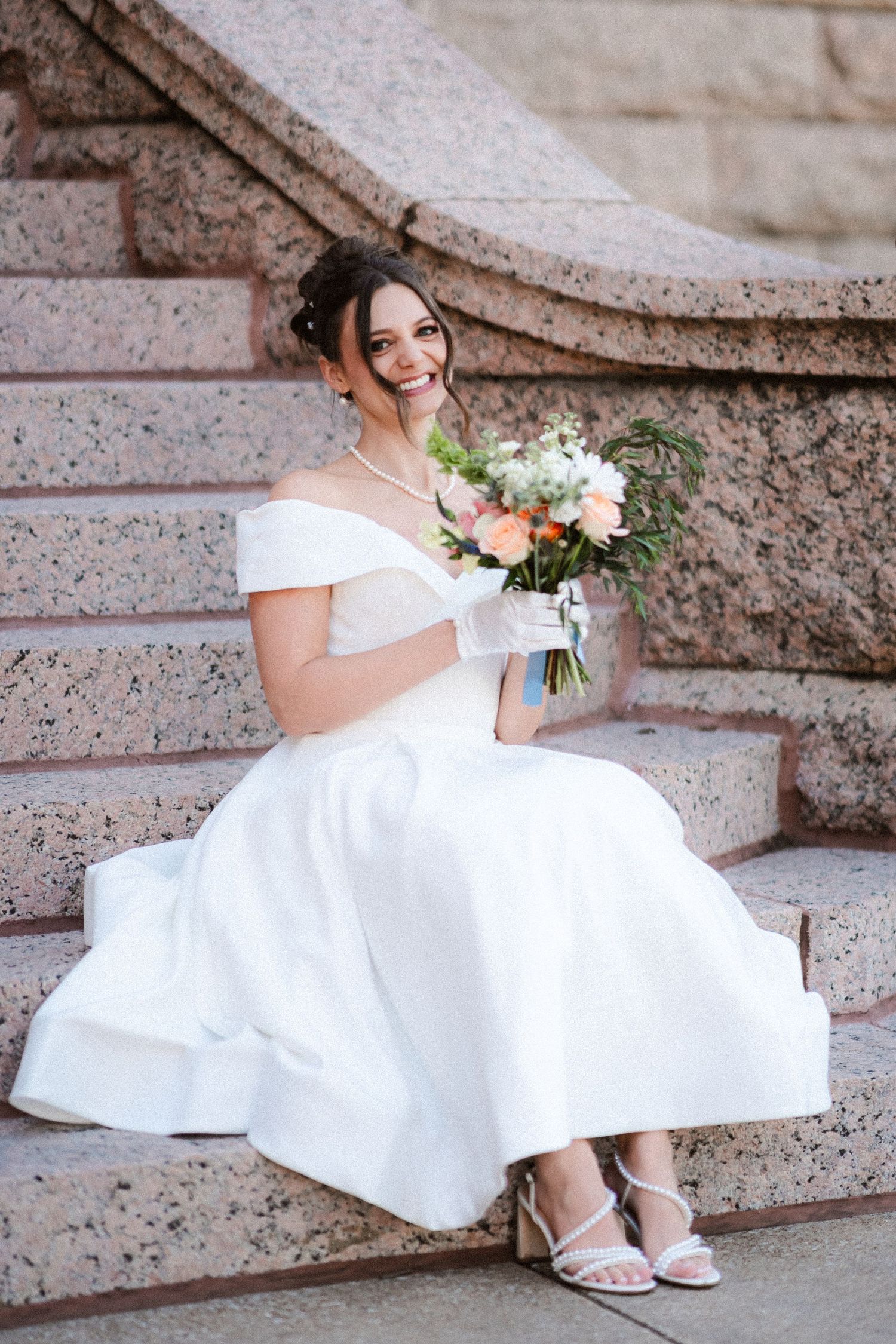 Bride sitting on stone steps holding a bouquet of pink and white flowers.