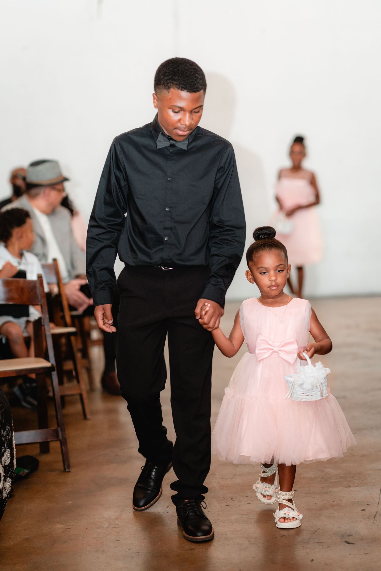 A young child in a pink tulle dress walking alongside an adult in formal black attire at an indoor event.