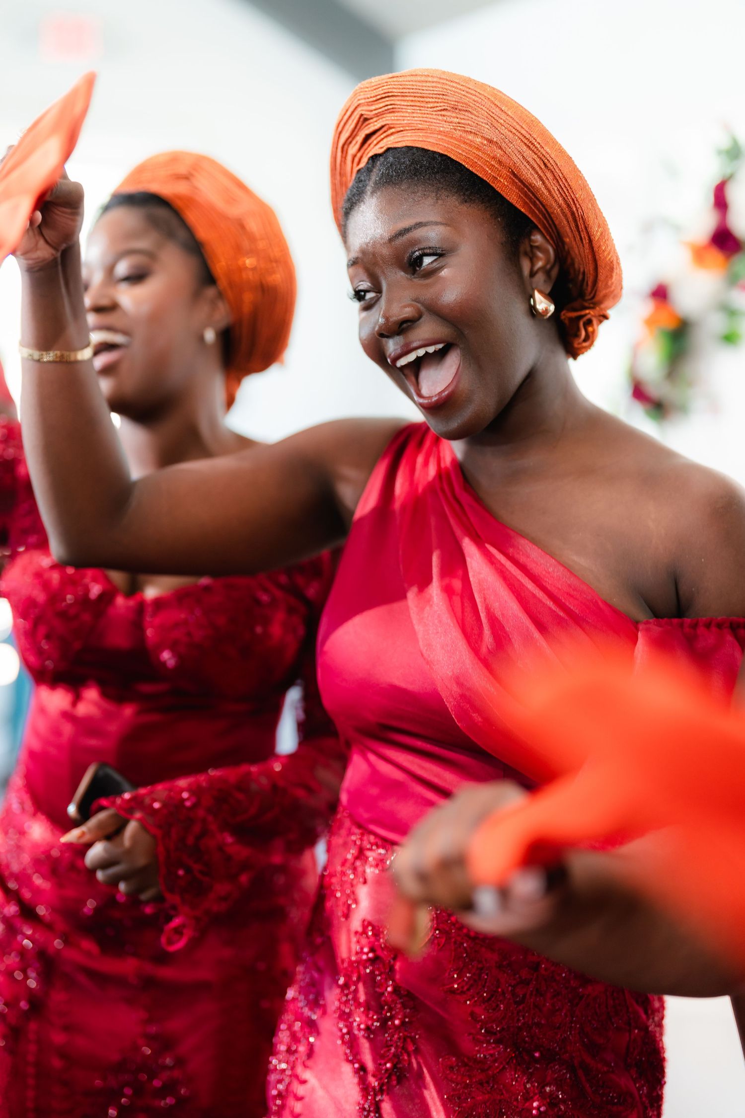 Attendees in red formal dresses and orange headwraps dance enthusiastically at a celebration.