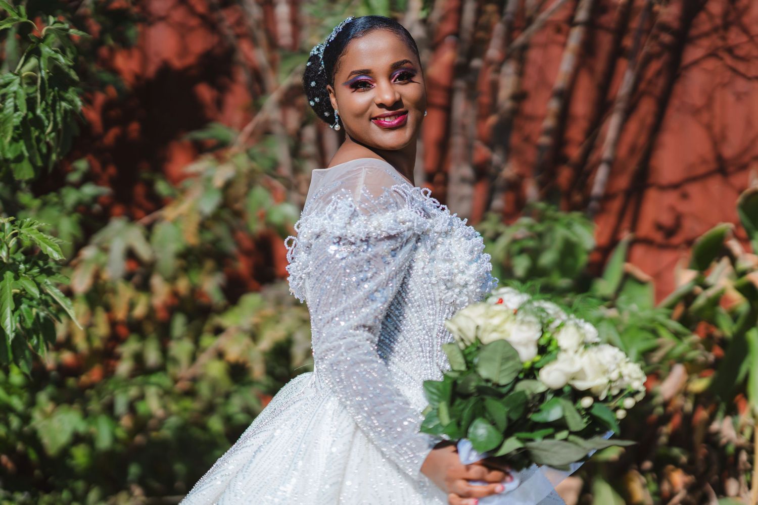 Elegant bride in a white lace ballgown poses with her bouquet of white flowers against a brick wall backdrop.