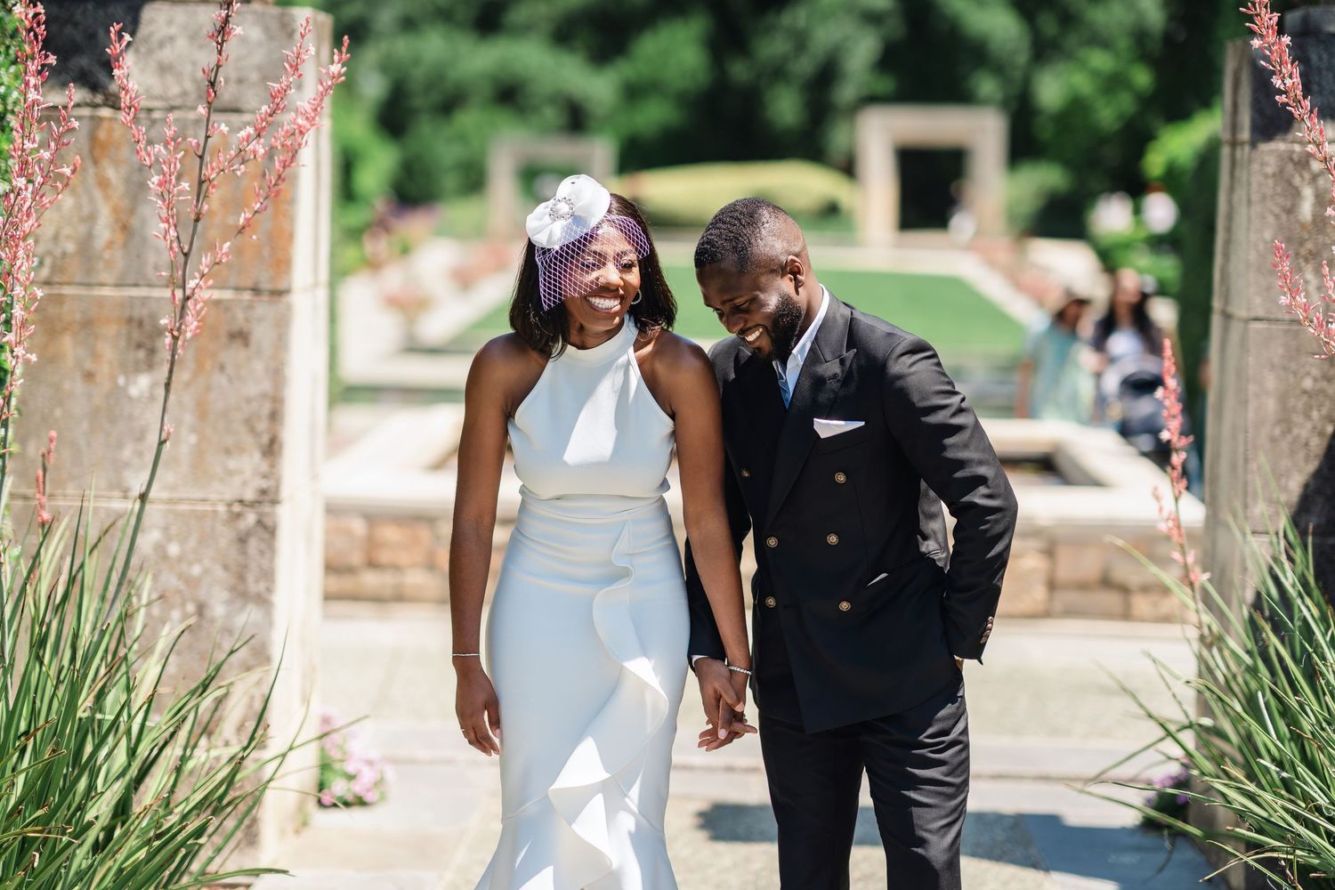 A couple in wedding attire walks together and laughs on a stone path surrounded by greenery and flowers.