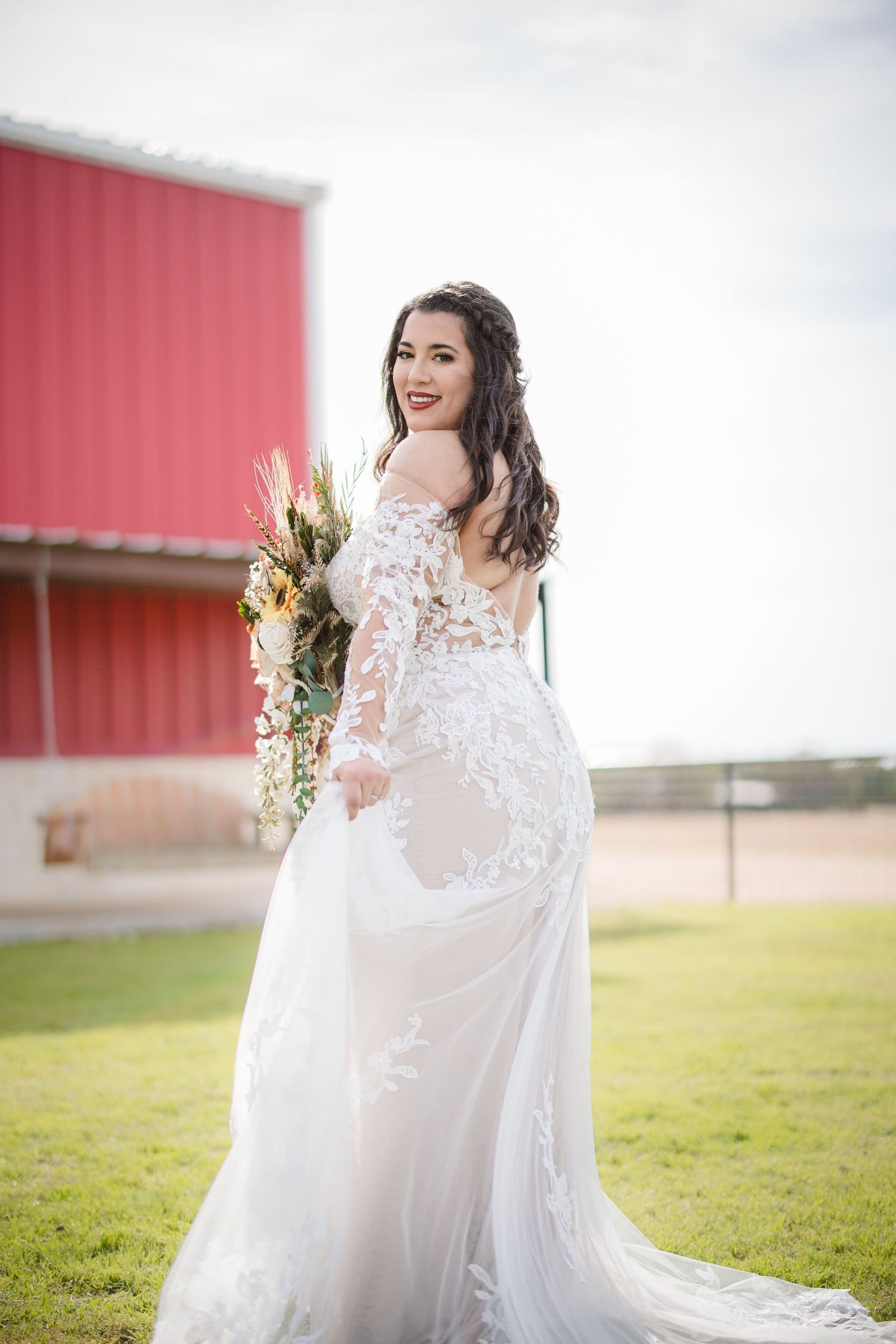 A bride in a delicate lace wedding gown twirls gracefully in an outdoor field with a red barn in the background.