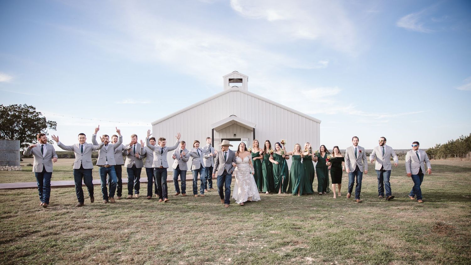 Wide panoramic shot of wedding party and guests gathered outside a white rural event venue.