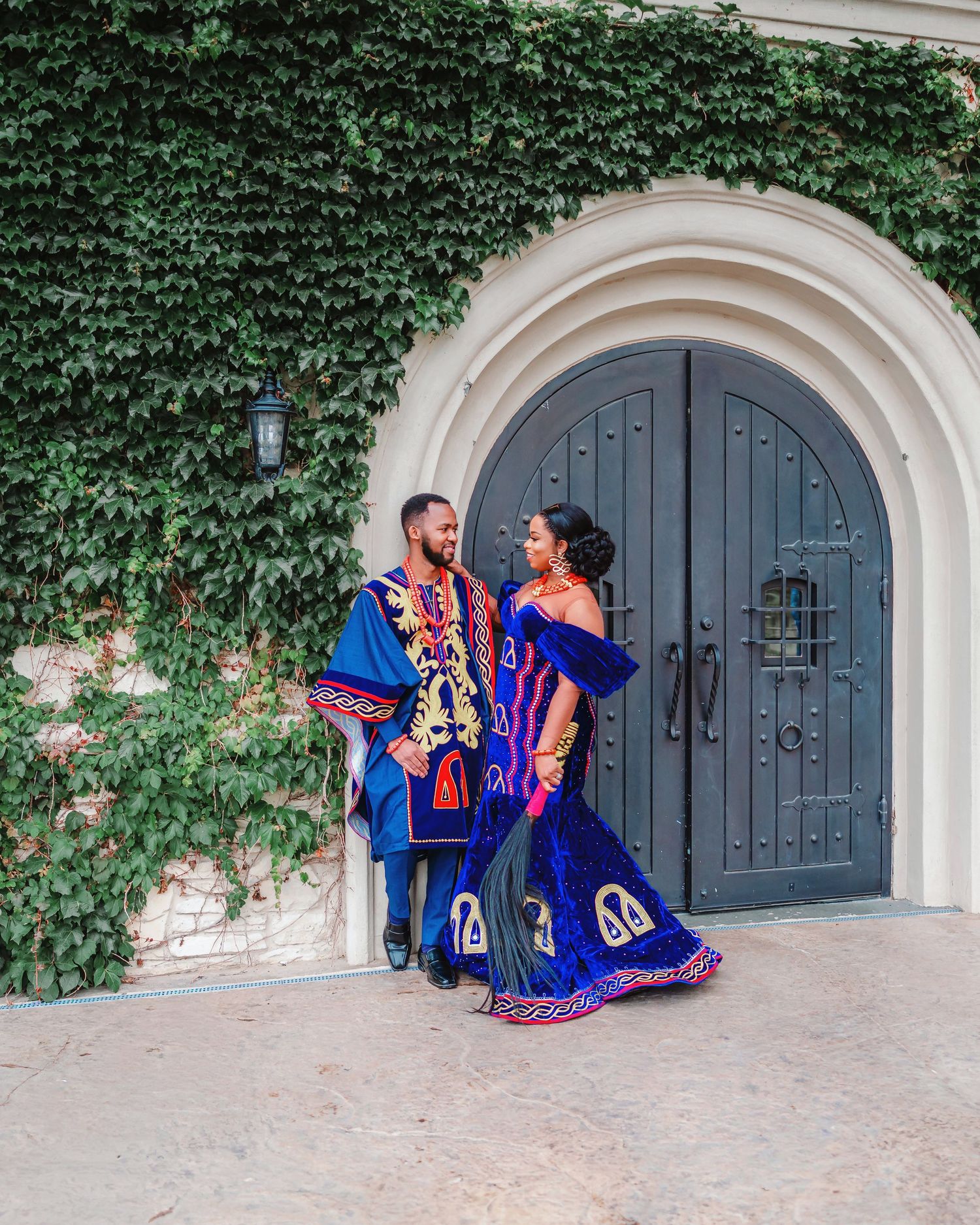 A couple in vibrant blue and orange traditional Mexican folk dresses pose in front of an ivy-covered stone wall.