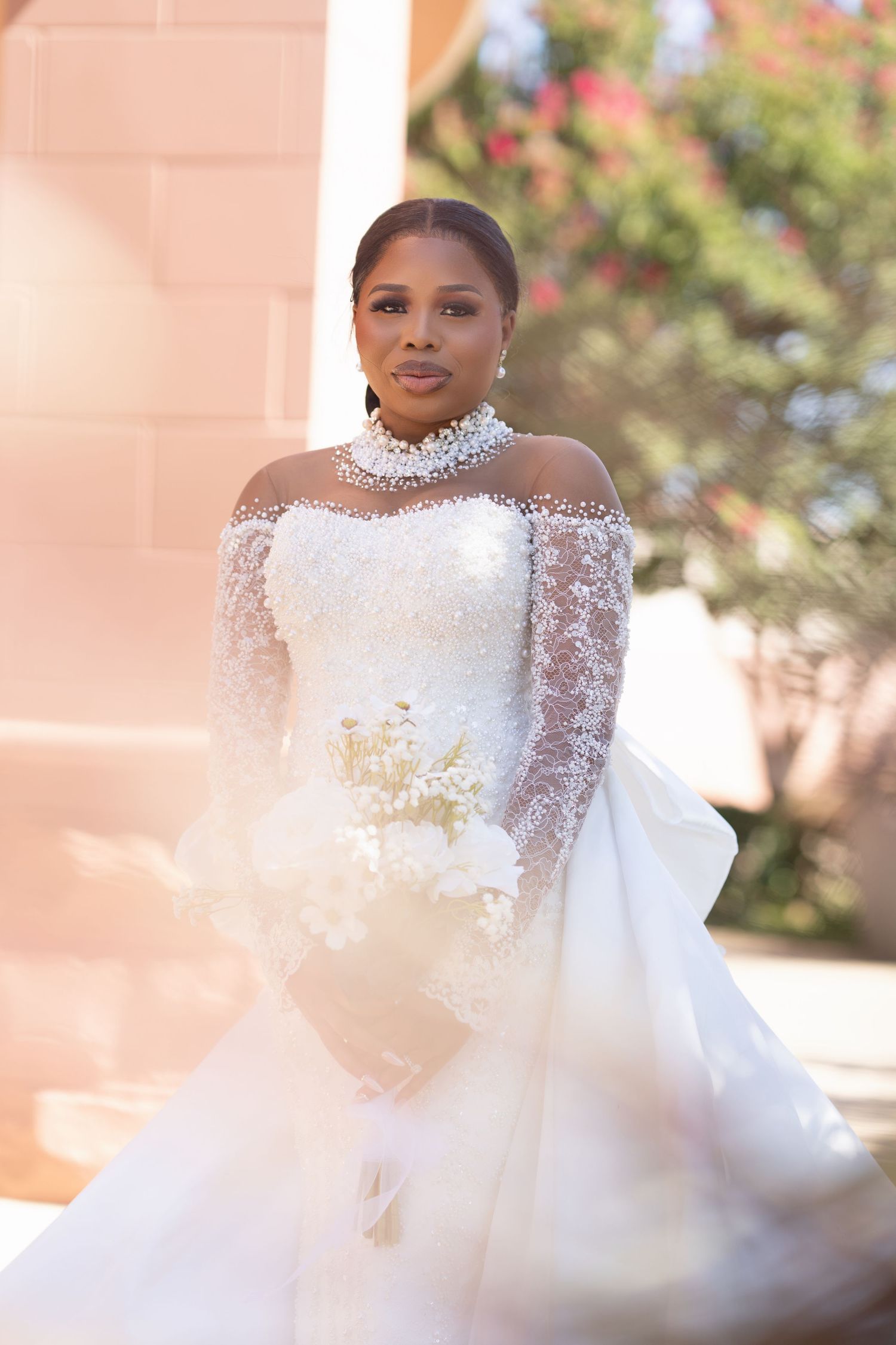 Elegant bride in a stunning white wedding gown with sheer lace sleeves and high neckline against a soft pink background.