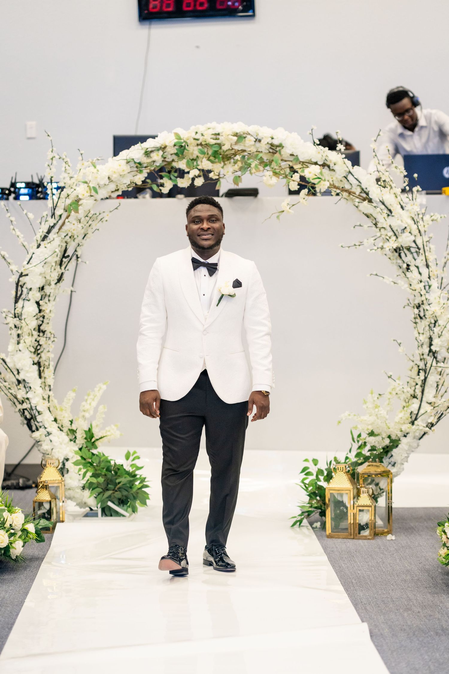 A groom in a white tuxedo jacket poses under a floral wedding arch.