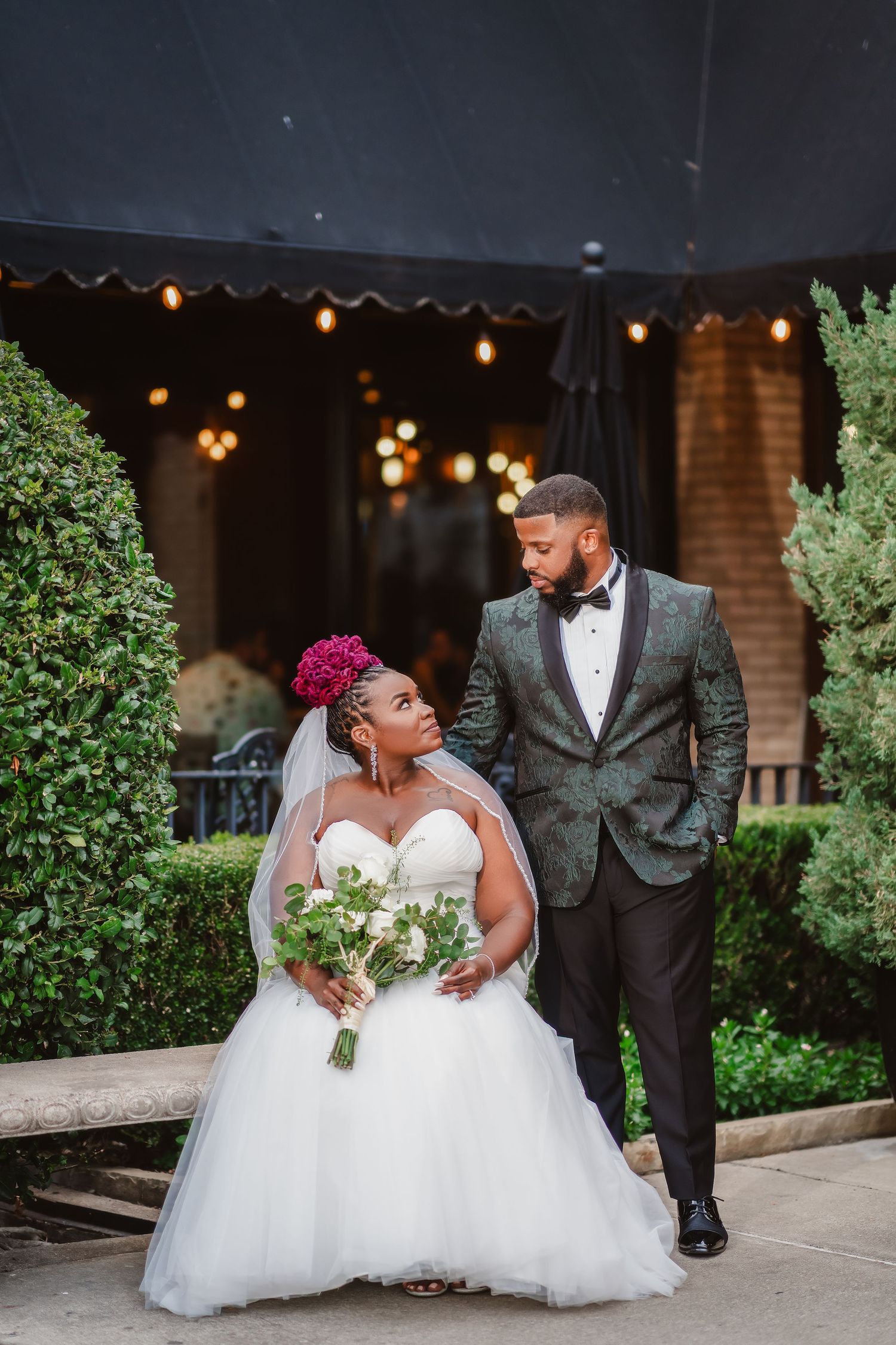 A romantic wedding couple poses outdoors at dusk with string lights and greenery creating an elegant ambiance.