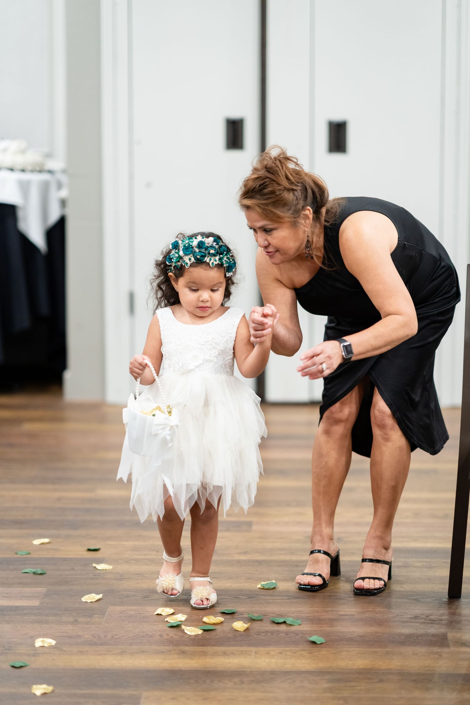 Playful moment as flower girl and woman in black dress pick up petals from wooden floor.