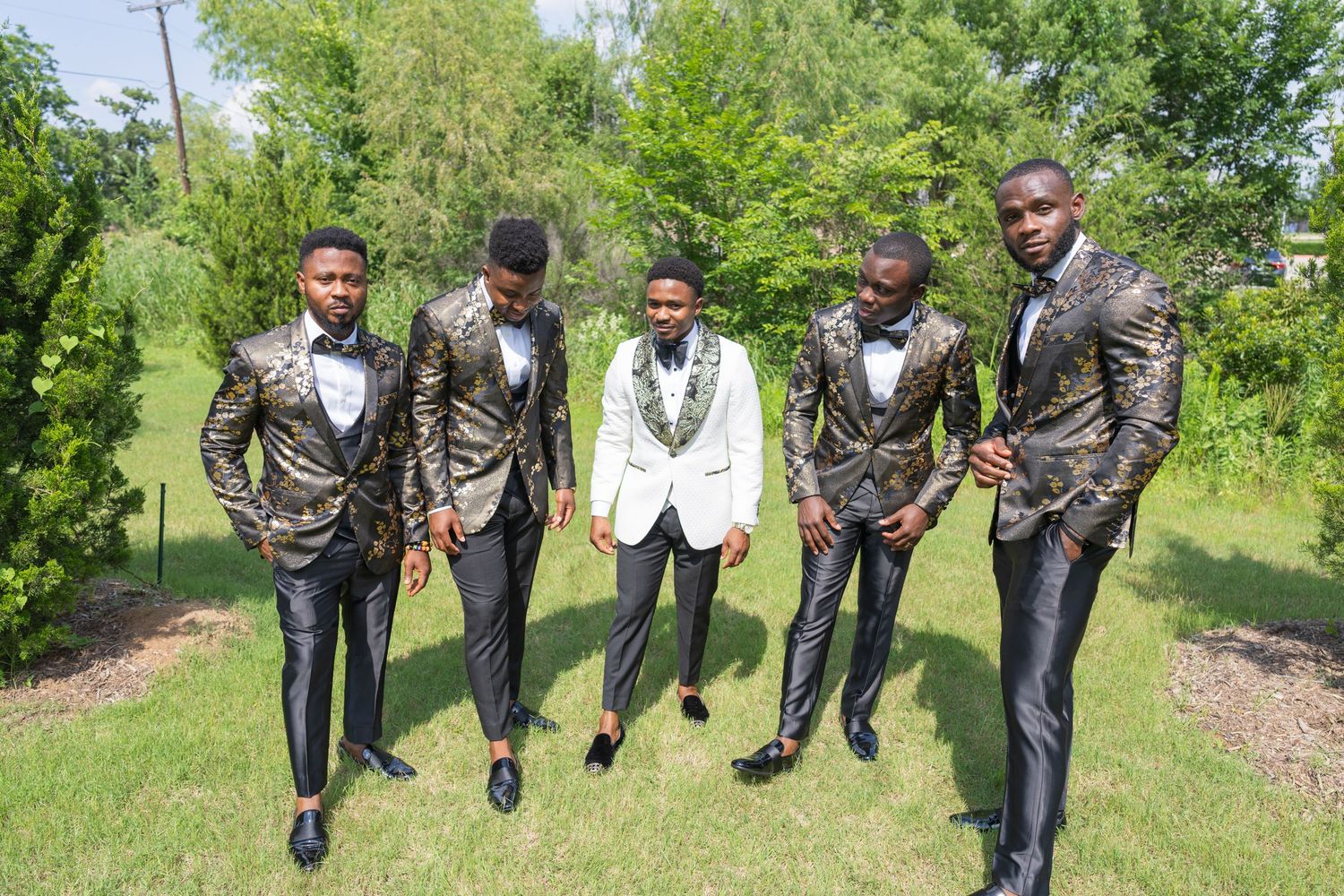 A group of groomsmen in stylish suits pose together outdoors on a sunny day with green foliage in the background.