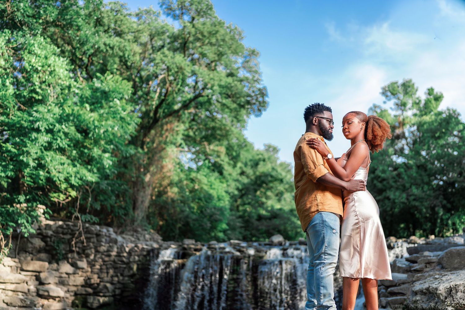 A couple shares a romantic moment on a park bench surrounded by lush green trees and stone walls.