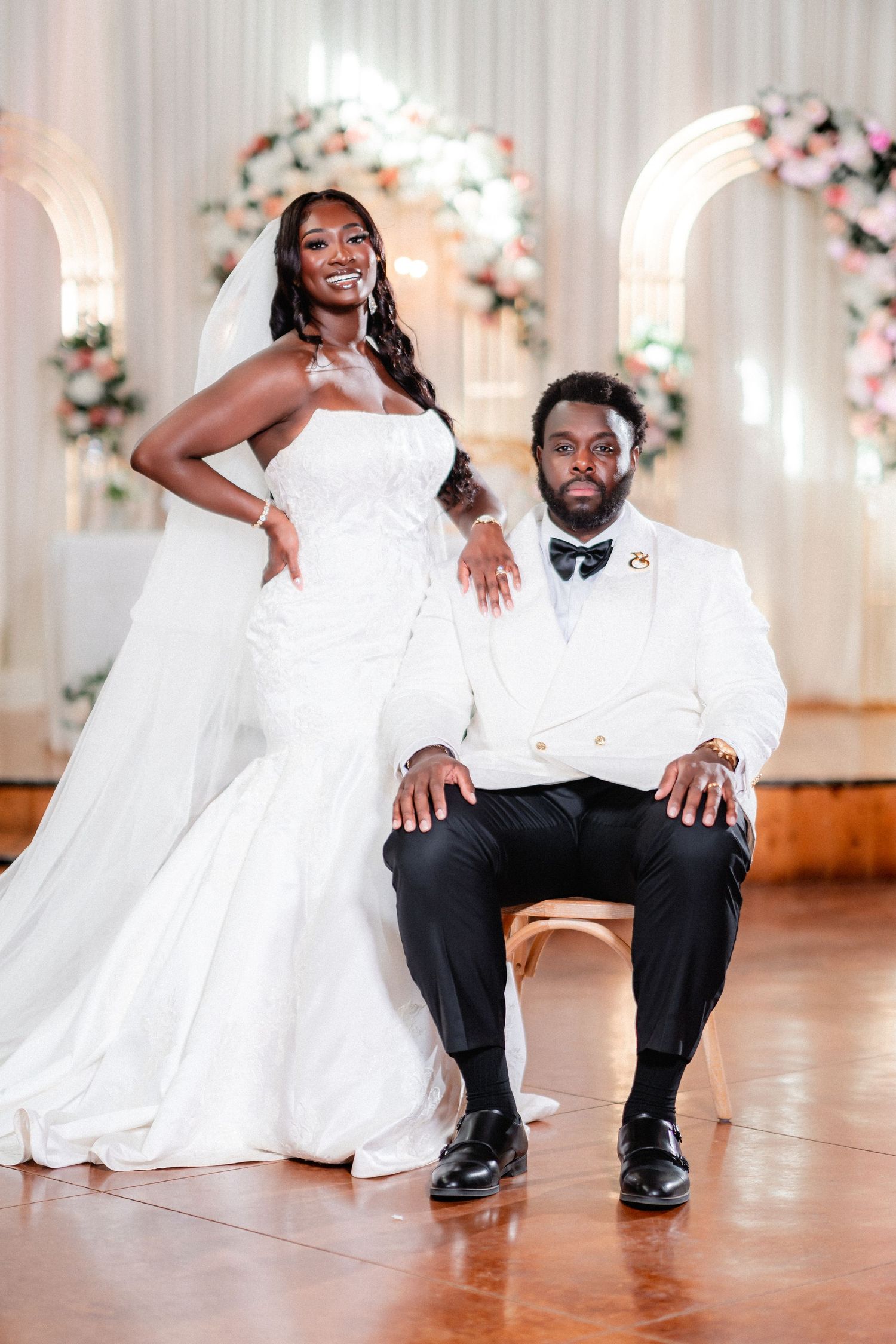 A newlywed couple poses playfully on a wooden dance floor with floral decorations and white curtains in the background.