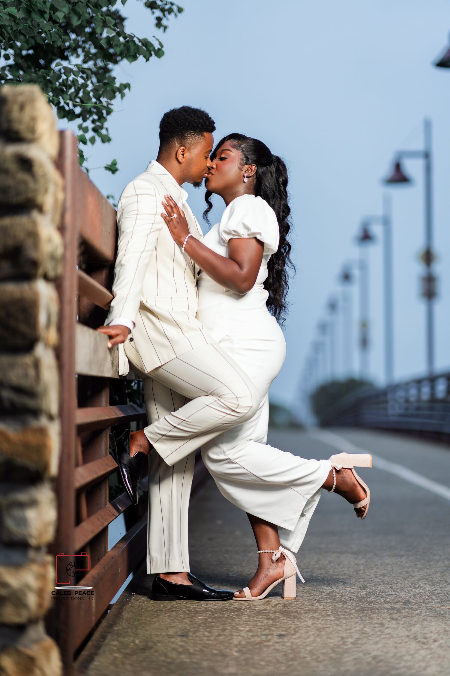 A romantic couple in white outfits share an intimate moment on a stone wall by a waterfront path.
