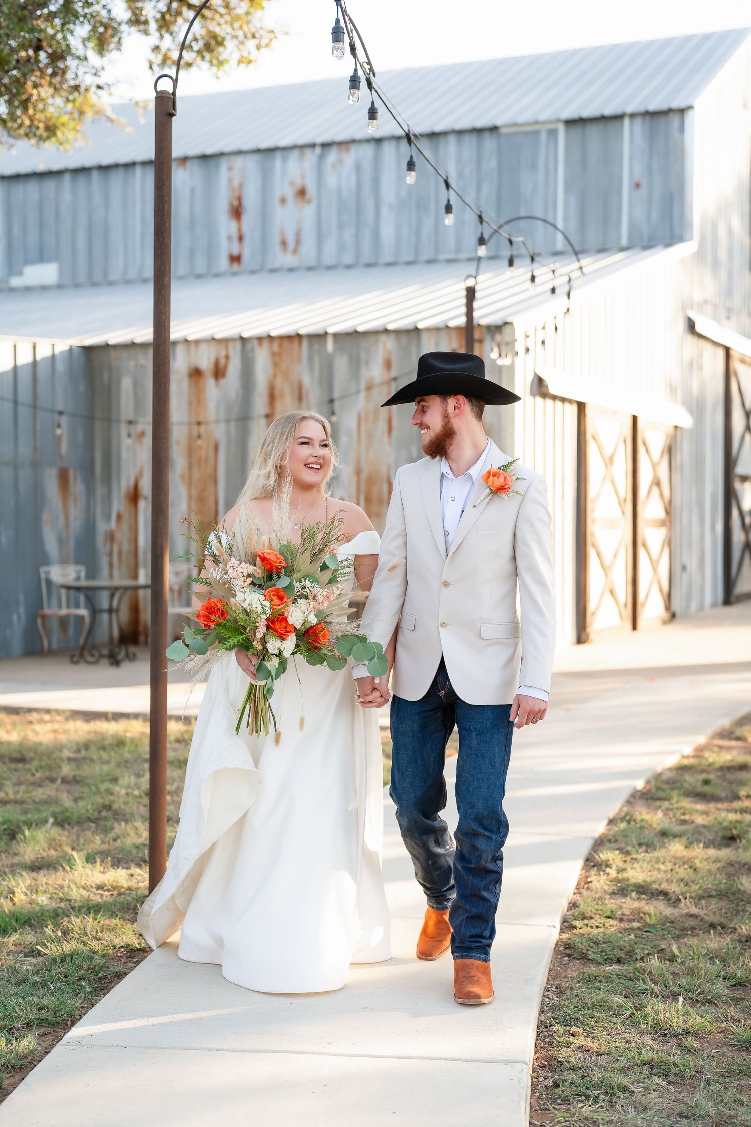 A rustic outdoor wedding scene with a couple walking along a path beside white metal buildings.
