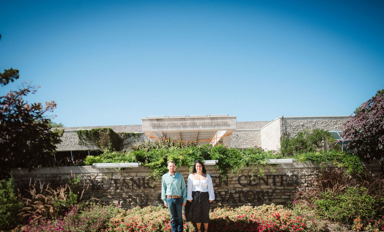 A couple stands together in front of stone garden walls on a clear blue day.