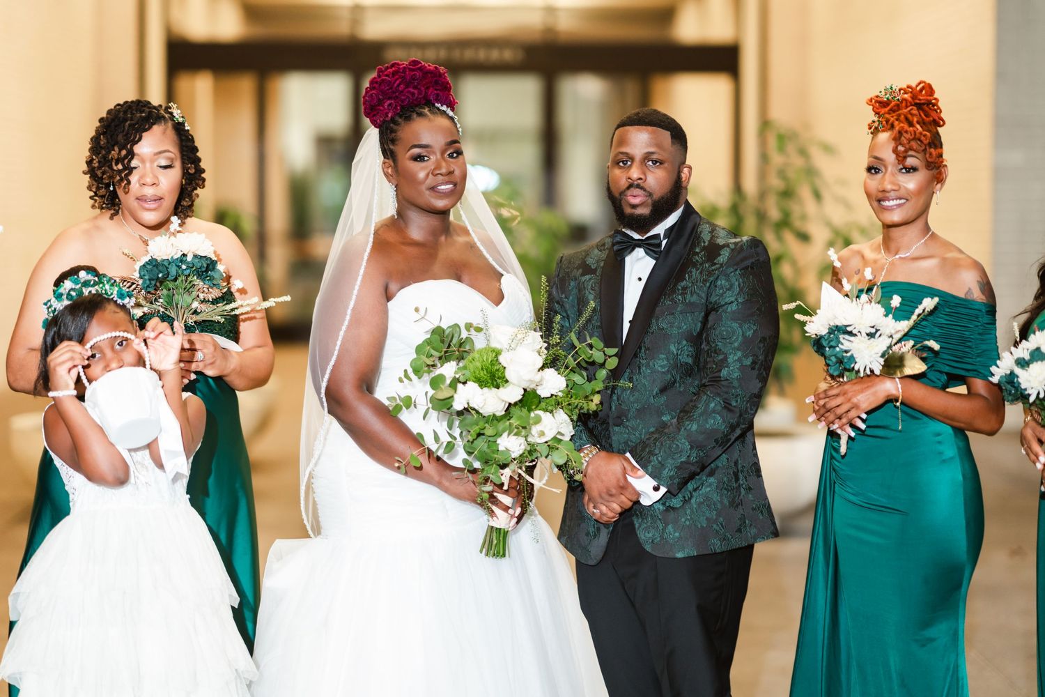 Wedding party in emerald green dresses and black tuxedos holding white floral bouquets.