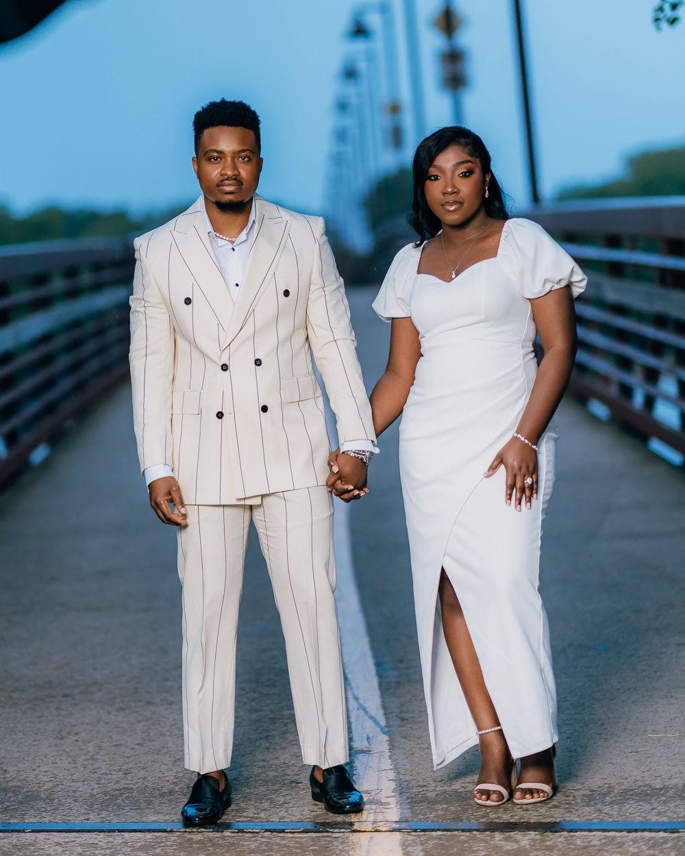 Couple in elegant white attire poses on a bridge during their engagement session.