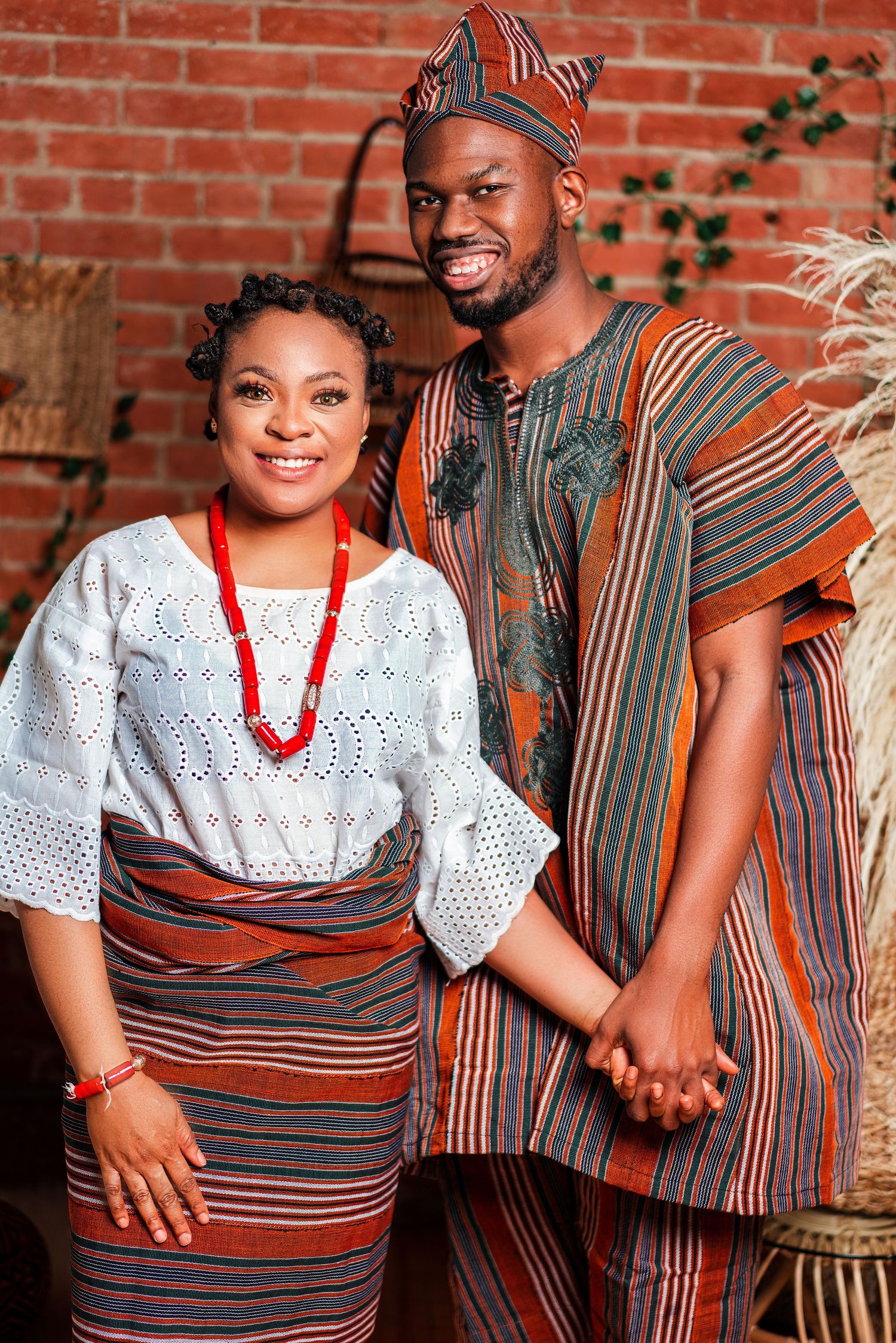 Couple in traditional African attire poses against brick wall wearing white lace and striped patterns.