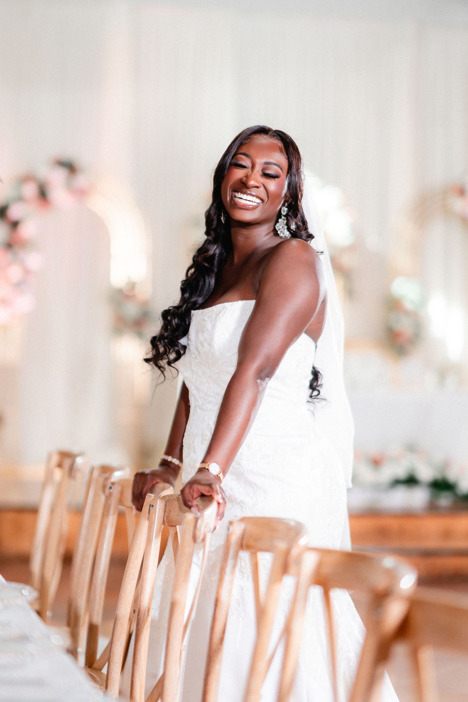 Radiant bride in white dress poses elegantly by wooden chairs in a bright indoor venue.