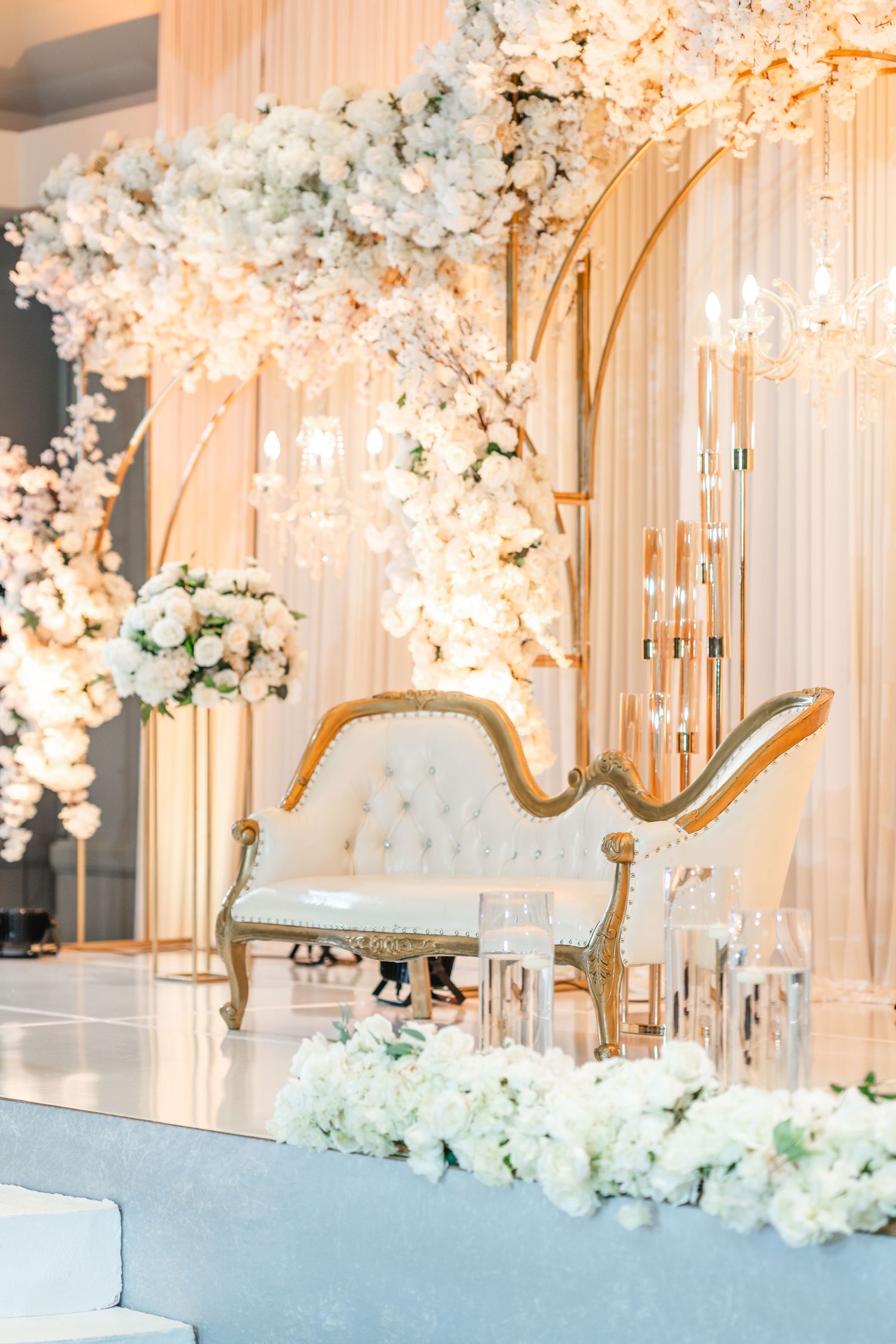 Elegant wedding reception setup with white floral arches and a luxurious tufted loveseat in gold and cream.