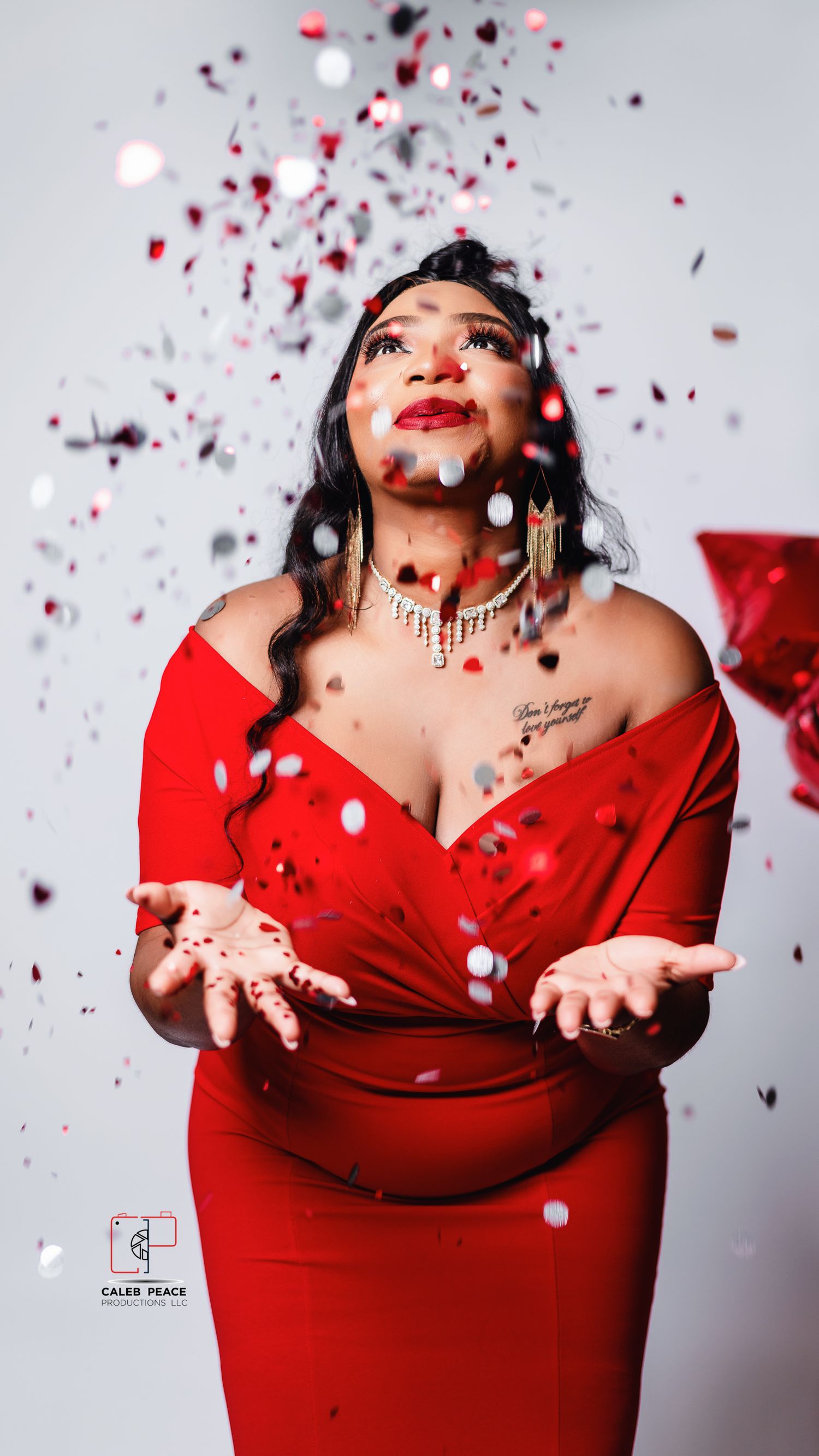 A glamorous portrait with red confetti falling around a person in an elegant red off-shoulder dress against a white backdrop.