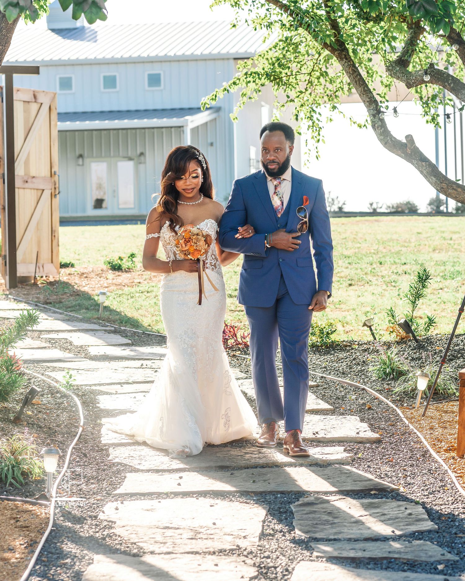 A couple in wedding attire walk together on a garden path, with the bride in white and groom in blue suit.
