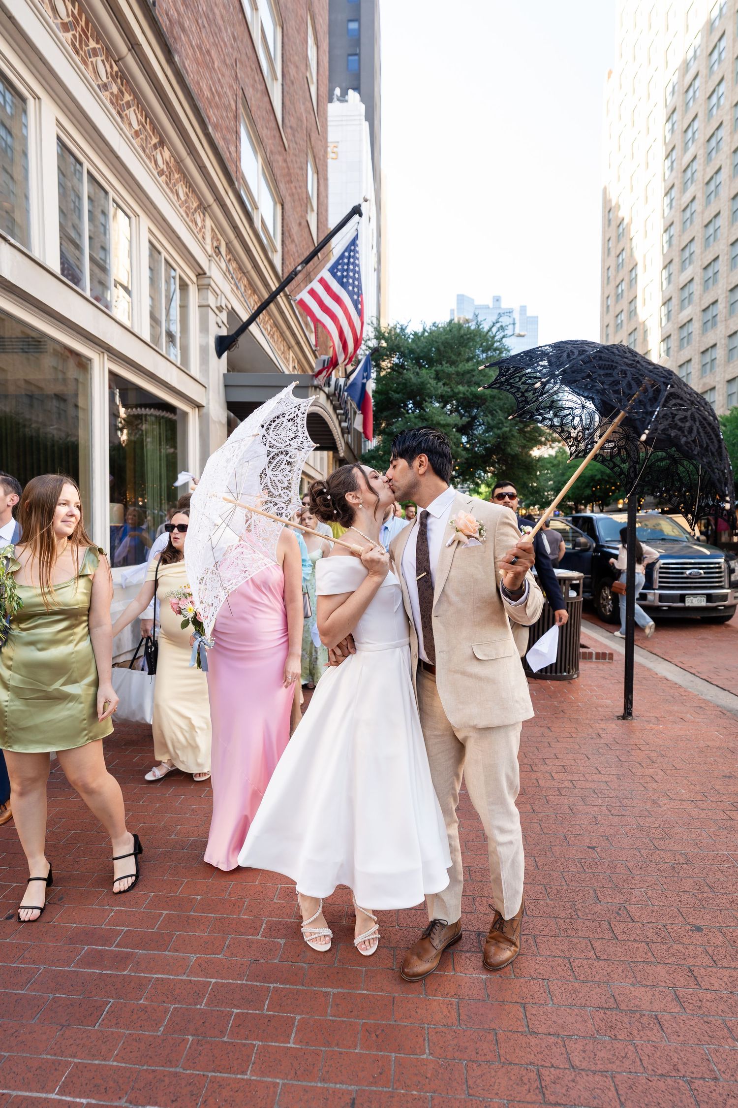 Wedding party walking together on brick sidewalk with American flag in urban setting.