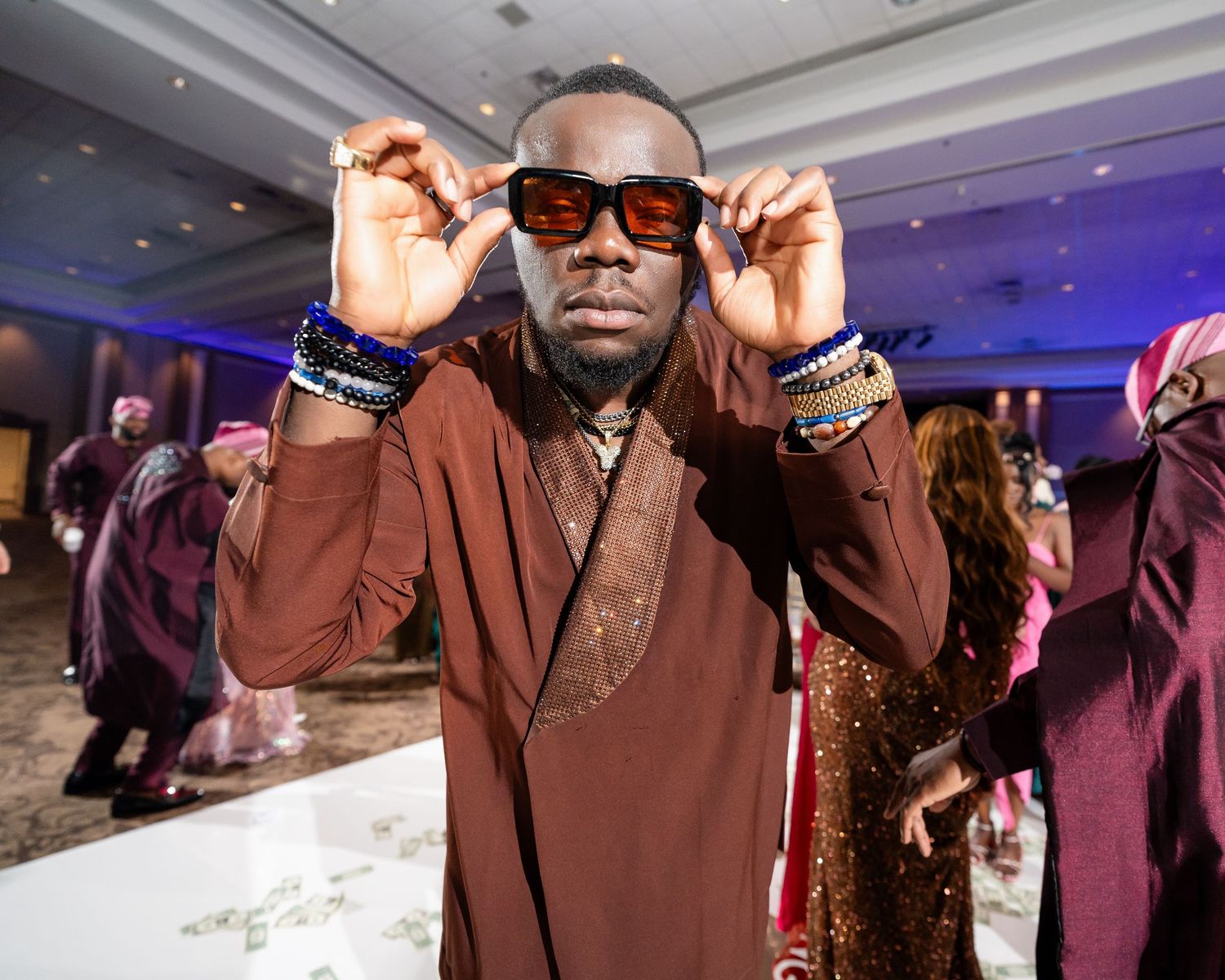 A person in a brown traditional outfit adjusts their sunglasses at an indoor event with purple lighting.