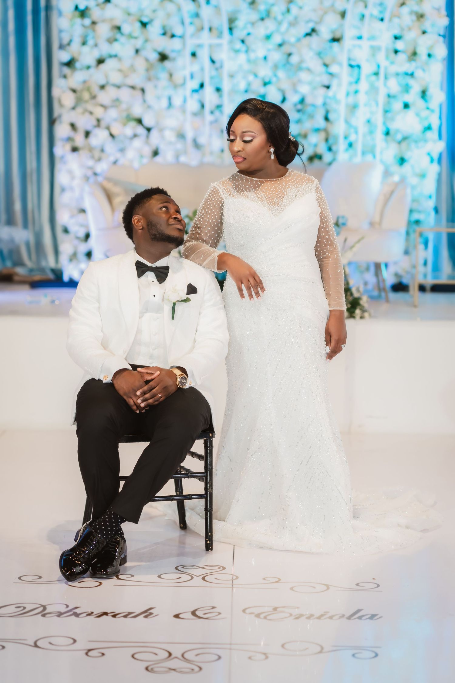 Elegant couple in white wedding attire pose together on a decorated floor with floral backdrop.