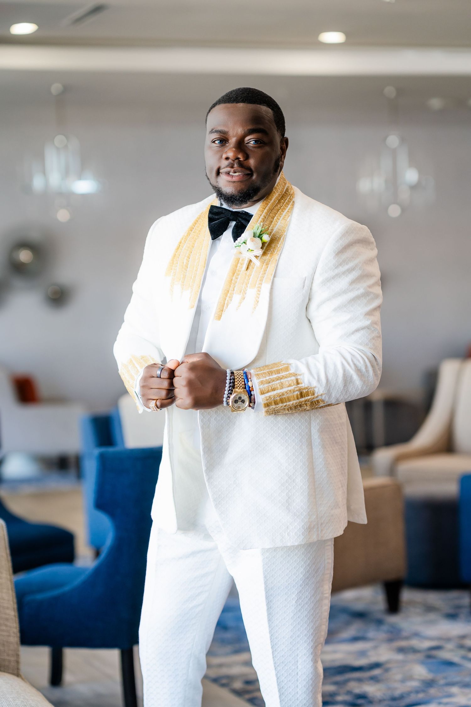 Groom poses confidently in crisp white tuxedo with gold accents and boutonniere against modern interior.