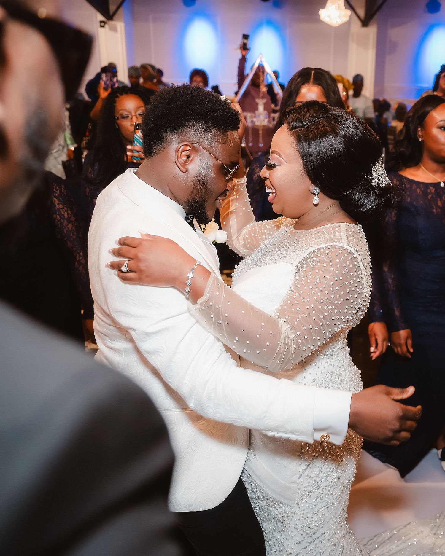 A couple shares a romantic dance at their wedding reception with blue lighting and guests in the background.