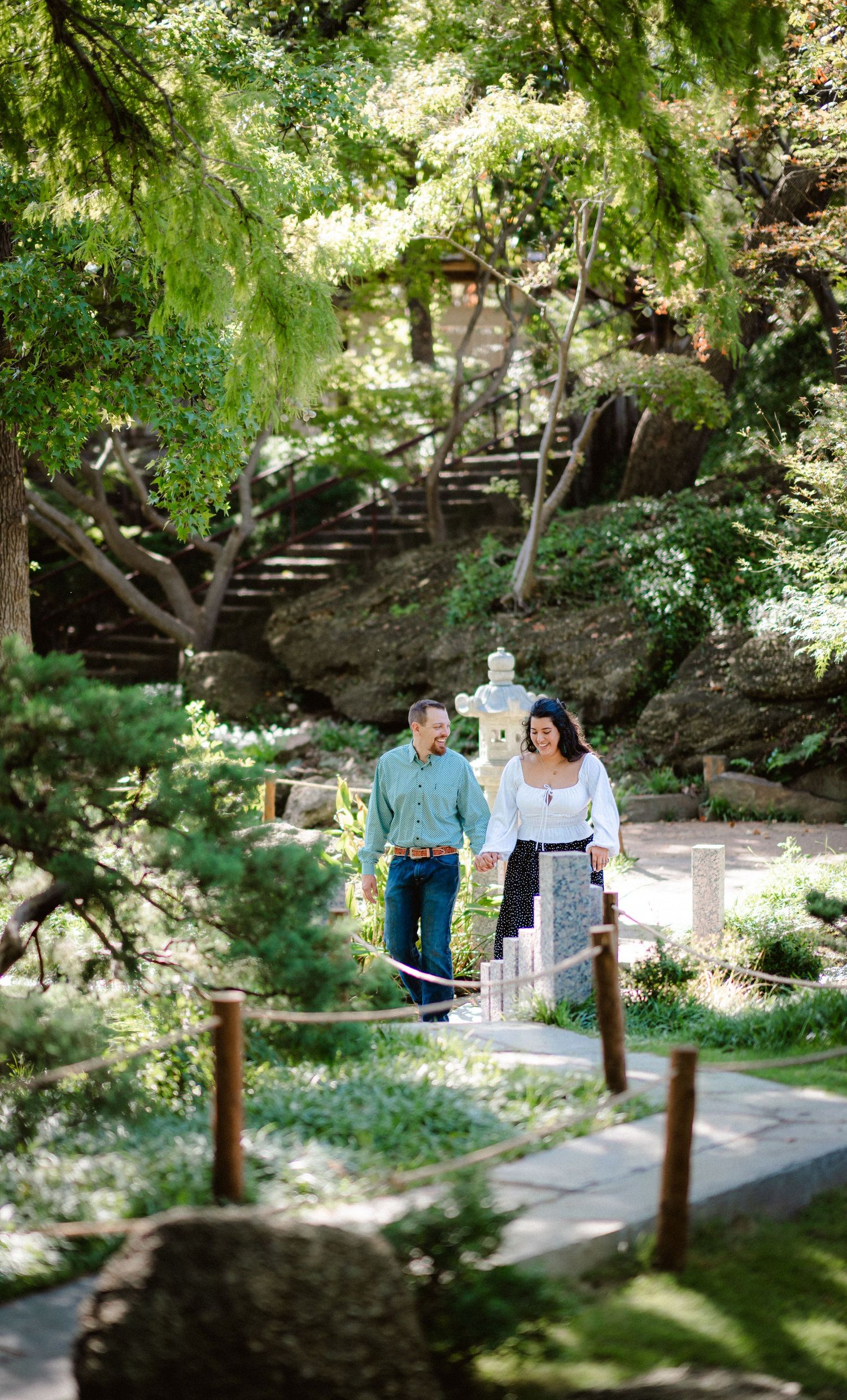 A couple strolls together along a garden path surrounded by lush greenery and wooden stairs.