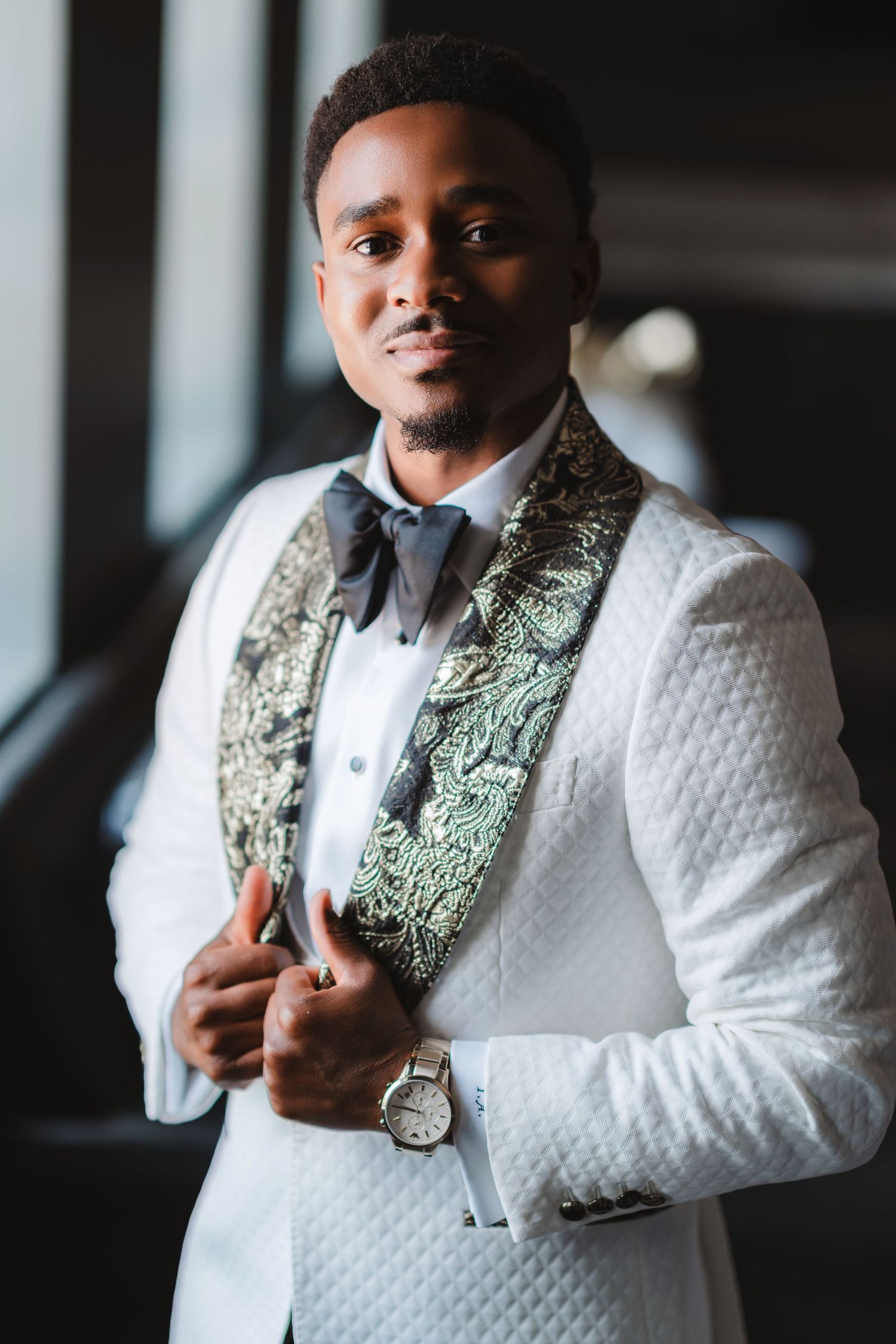 Groom wearing white textured jacket with ornate black and gold lapel detail adjusts his watch.