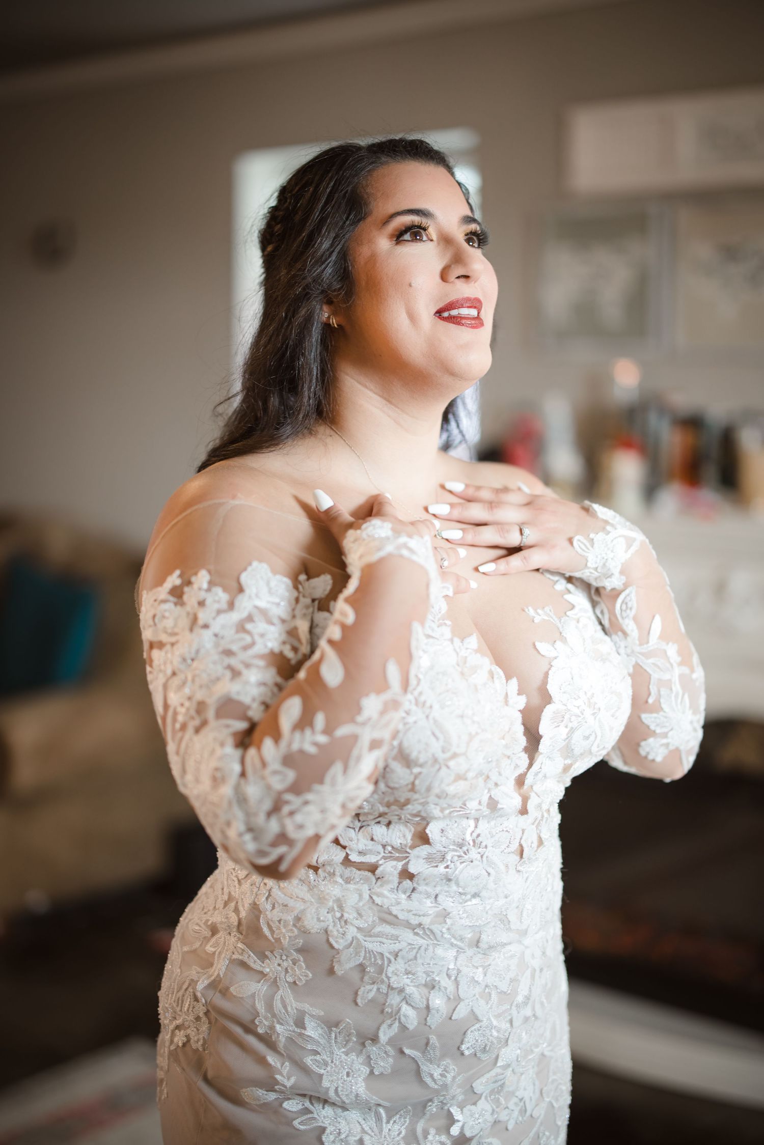 A bride wearing an elegant long-sleeved lace gown clasps her hands near her heart with a joyful expression.