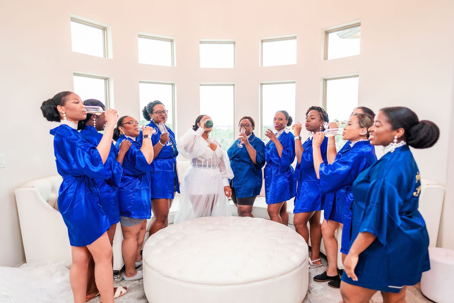A bridal party wearing matching royal blue robes gathers in a circle around a white ottoman during wedding preparations.