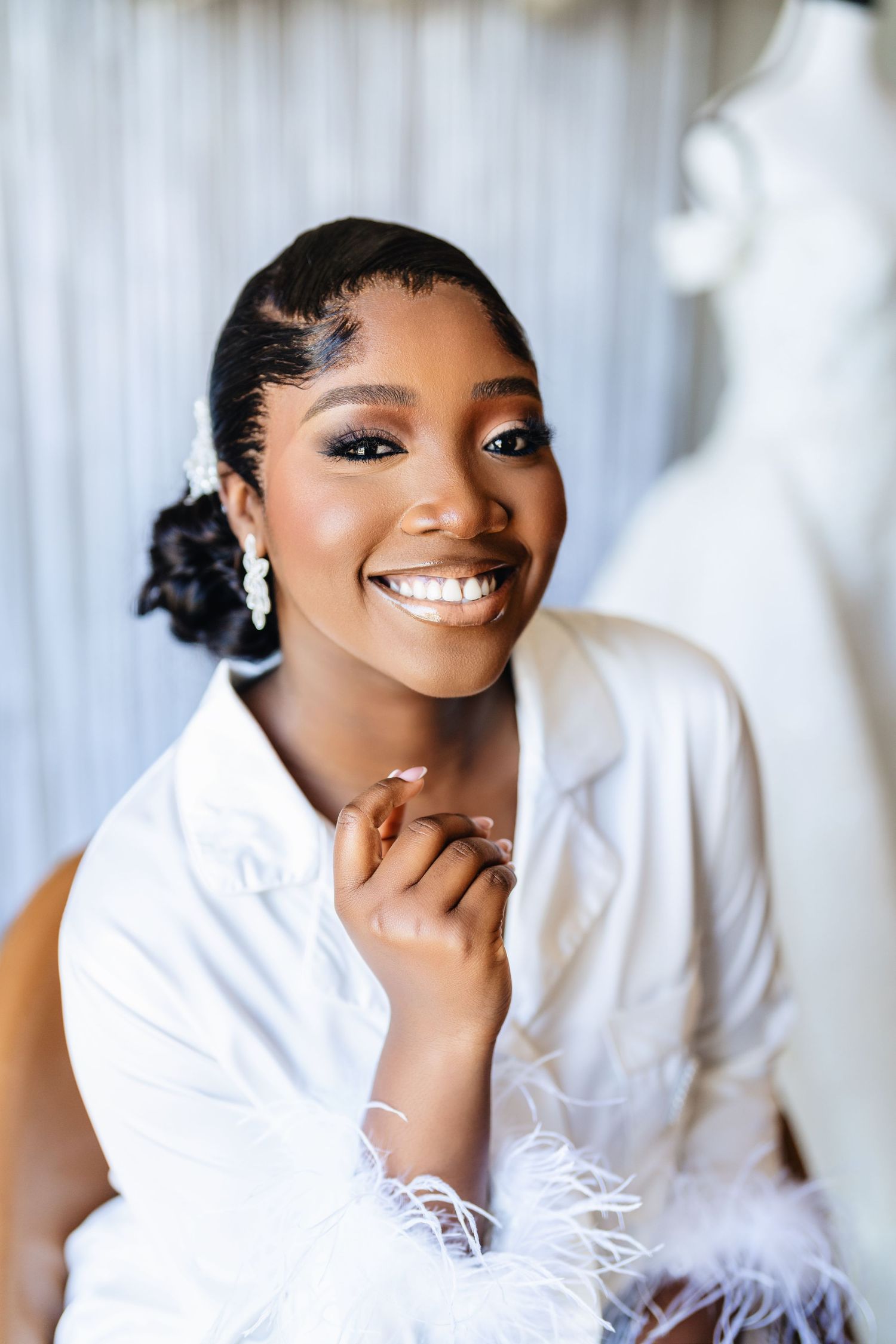 A bride in a white feathered robe poses with a radiant smile against a light backdrop.