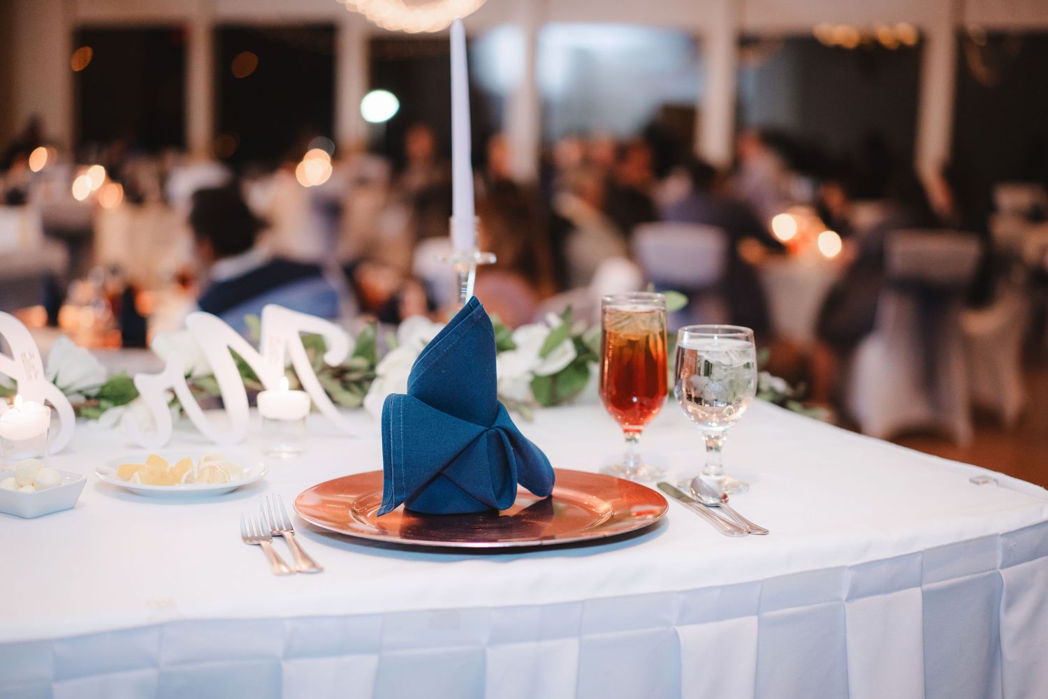Elegant wedding reception table settings with blue napkins and glowing candle centerpieces in a dimly lit venue.