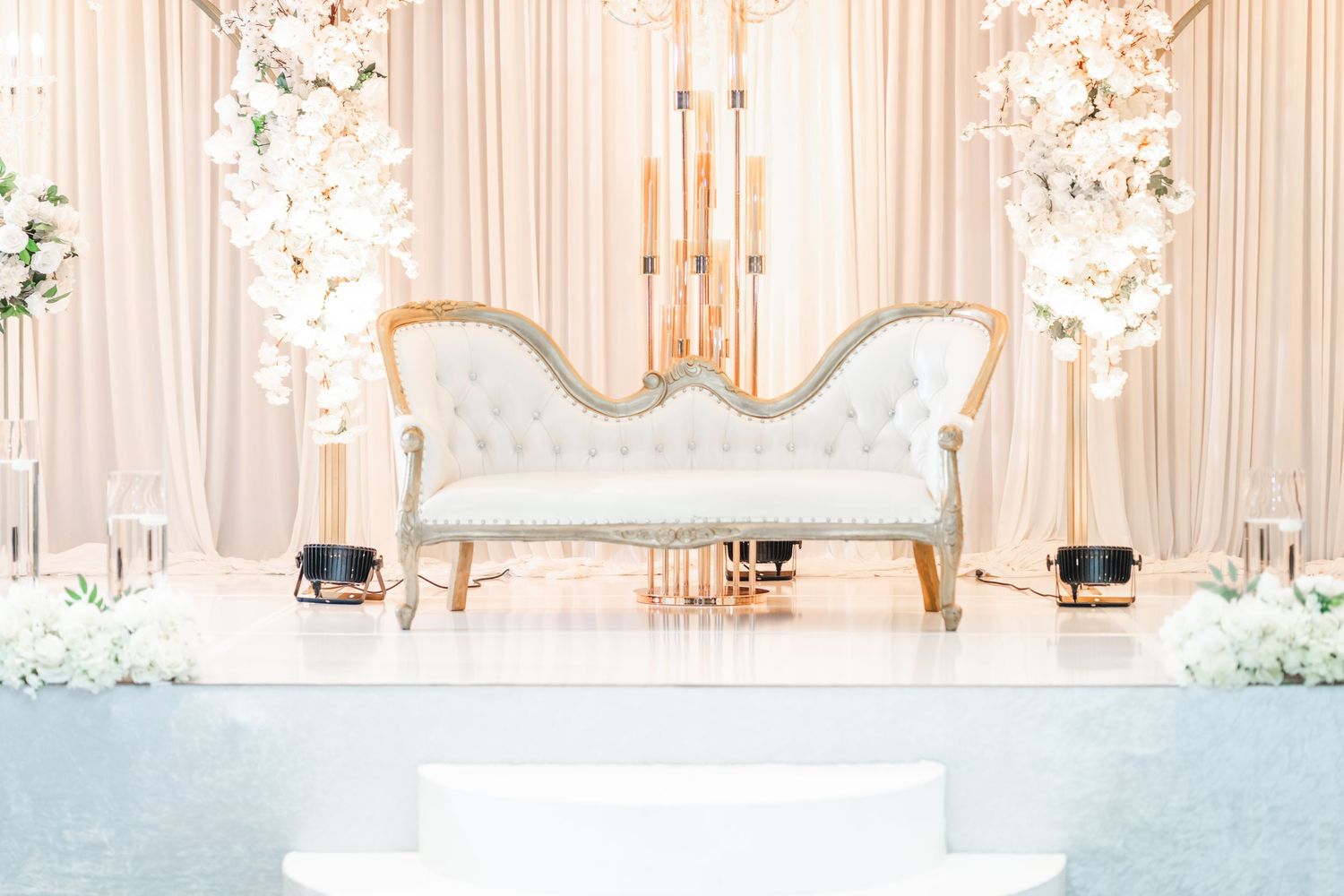 Elegant wedding reception venue with white drapery, floral arrangements and crystal chandeliers.