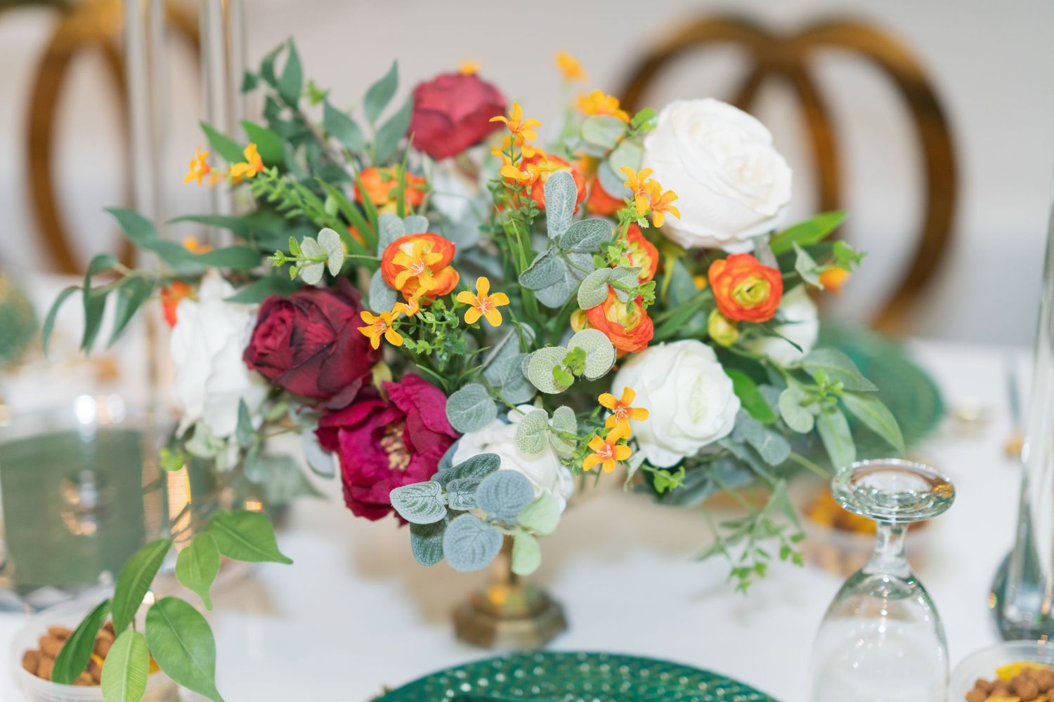 Colorful wedding centerpiece with burgundy roses, white peonies and orange flowers in gold vase.