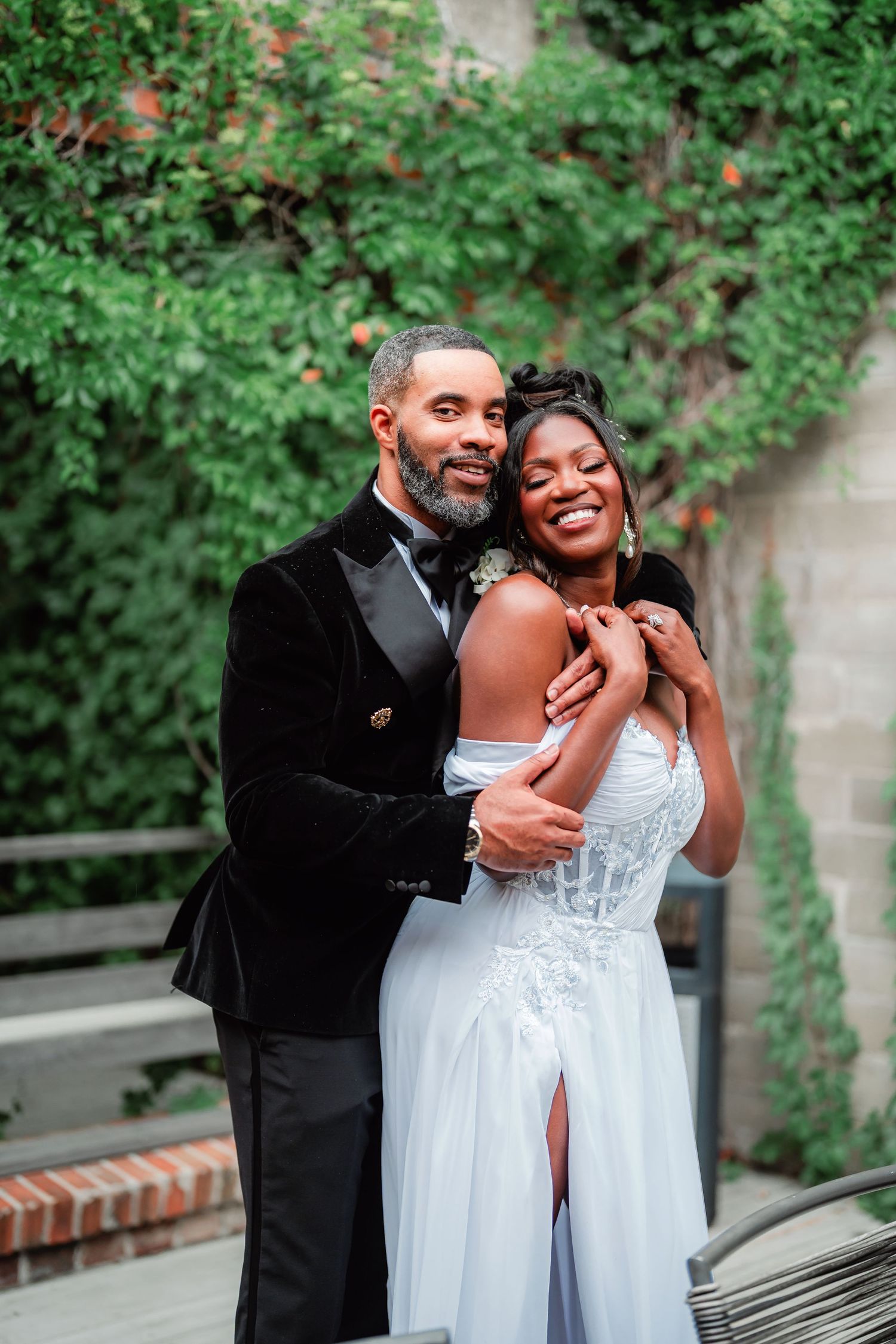 A joyful wedding couple embracing outdoors against lush green foliage.