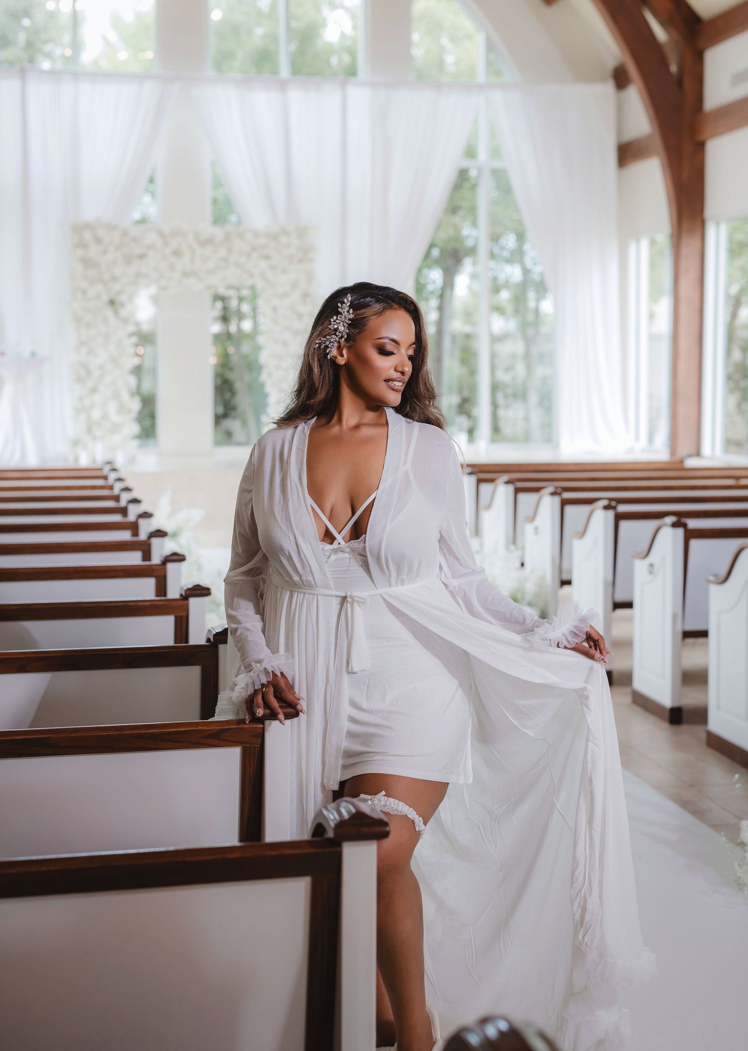 Bride in romantic white robe poses in chapel with wooden pews and natural lighting.