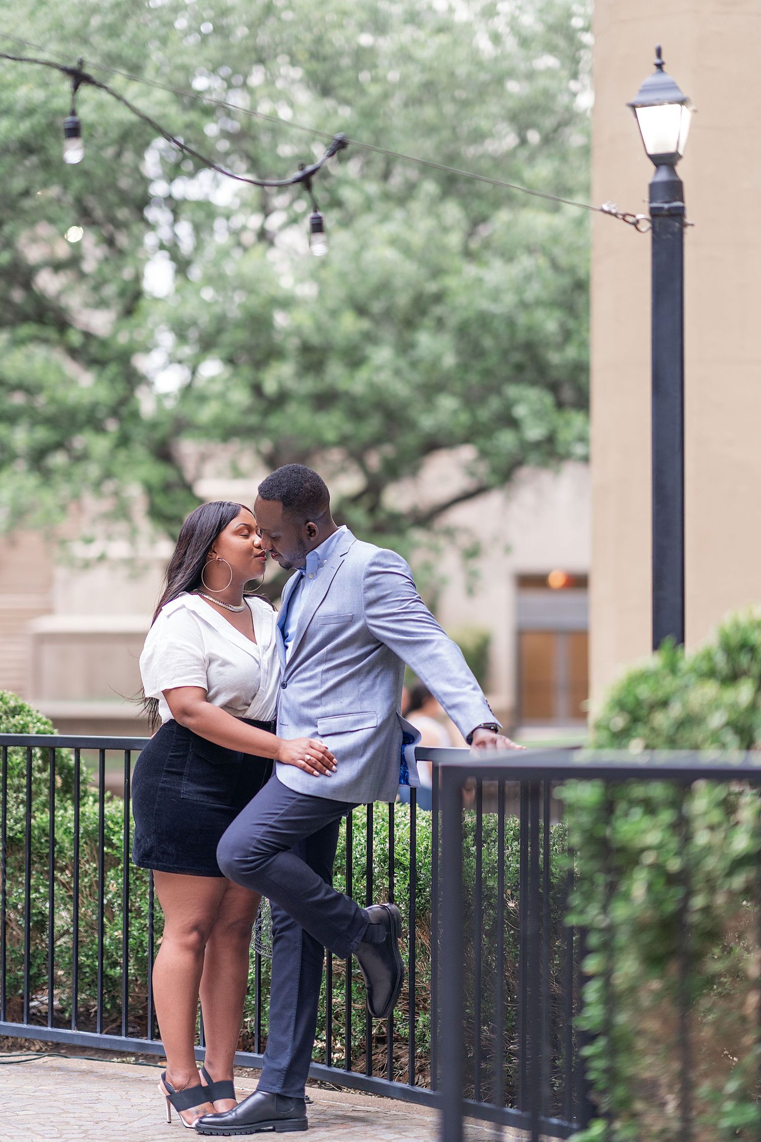 A romantic couple shares a tender moment by a railing with decorative lamp posts and greenery in the background.