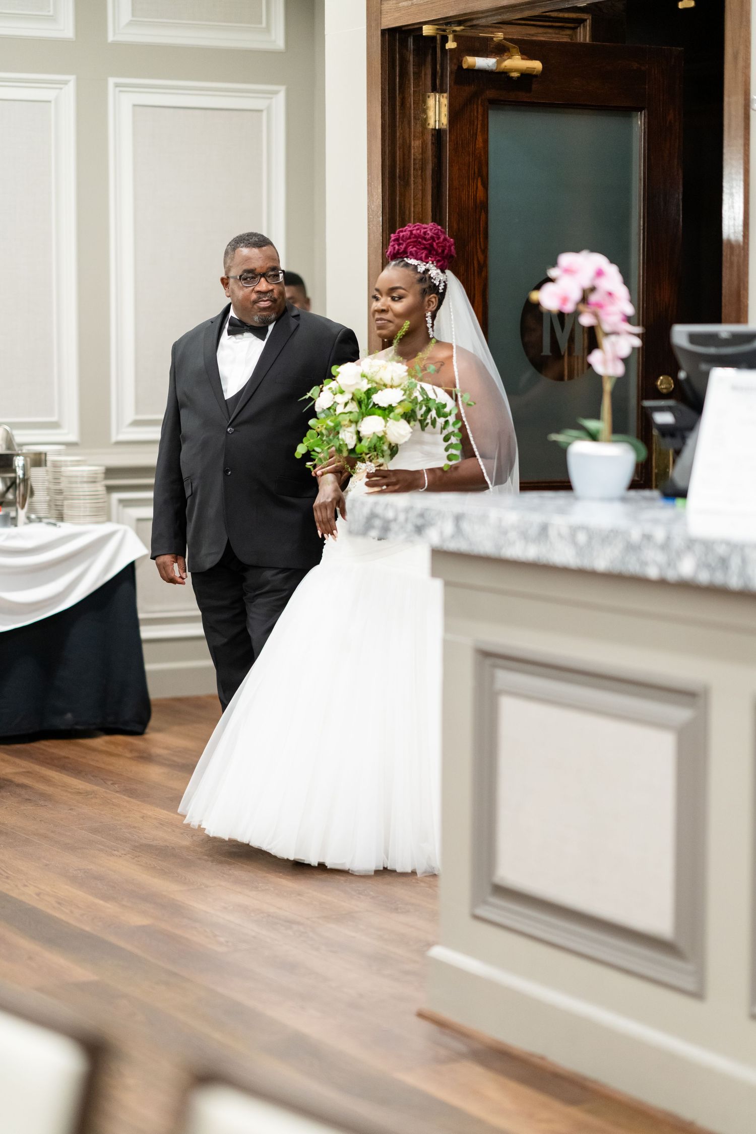 Wedding party members walk through elegant venue with white wainscoting and hardwood floors.